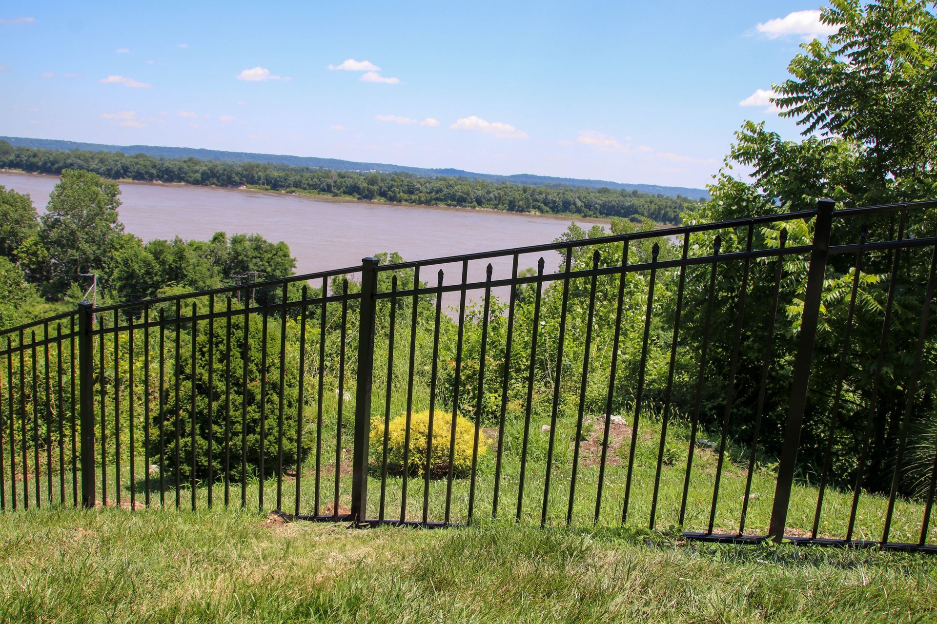 Black metal fence overlooking a river and treeline on a sunny day.