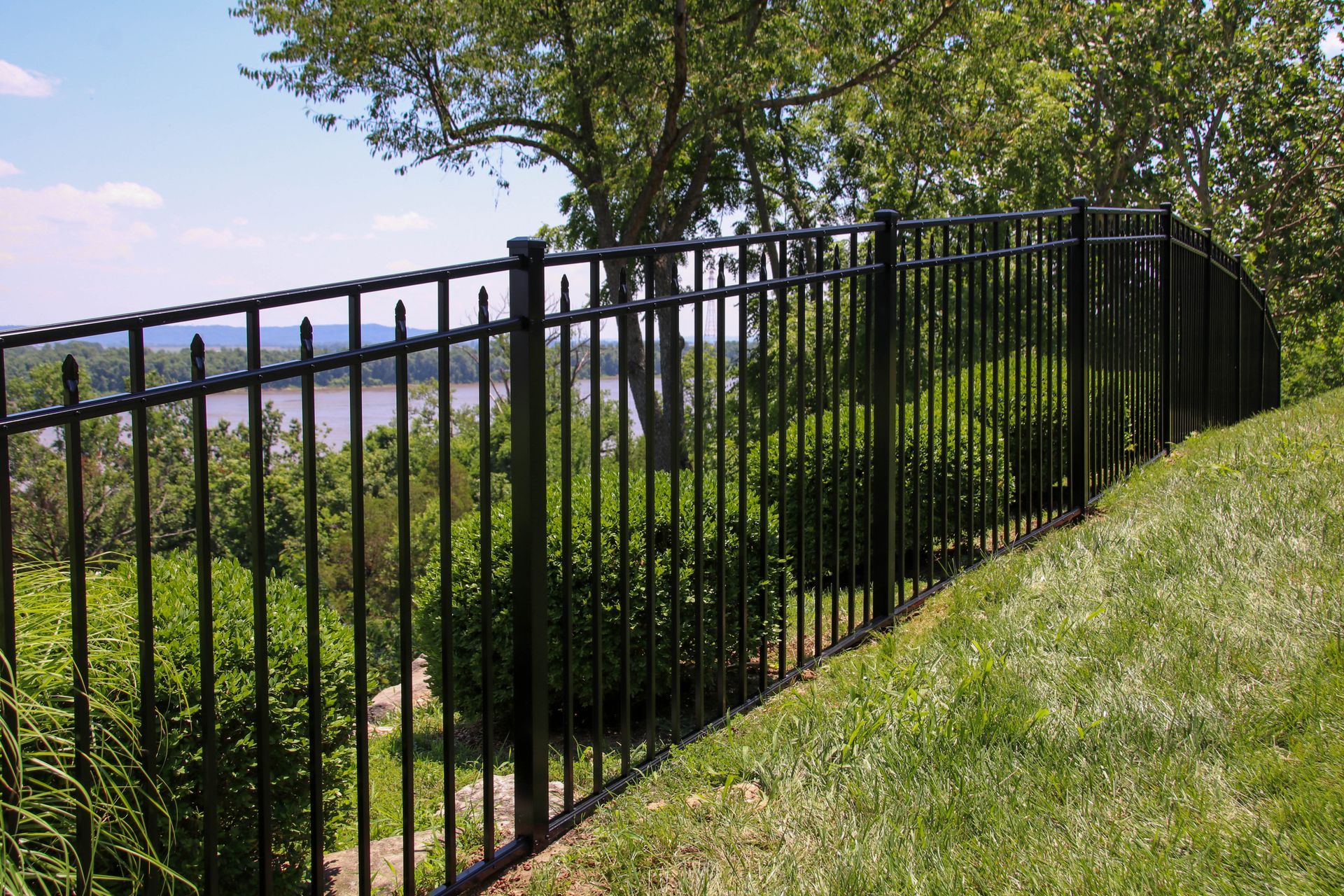 Black metal fence on a grassy hill, overlooking a river and trees under a sunny sky.