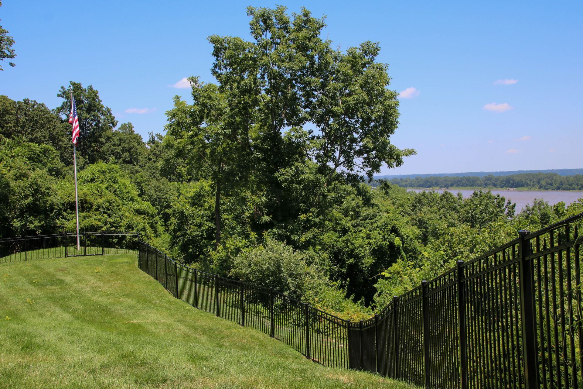 Green grassy hill with black fence, trees, American flag, and water view under blue sky.