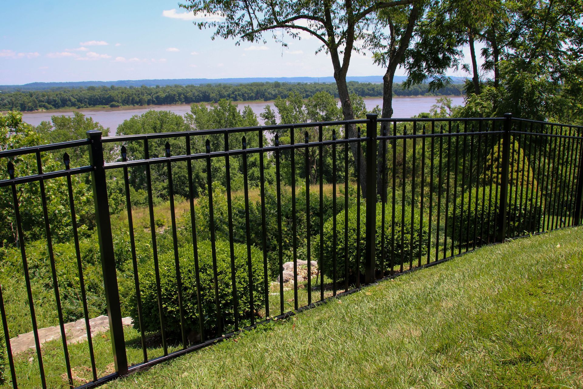 Black metal fence on a grassy hill overlooking a river and treeline.