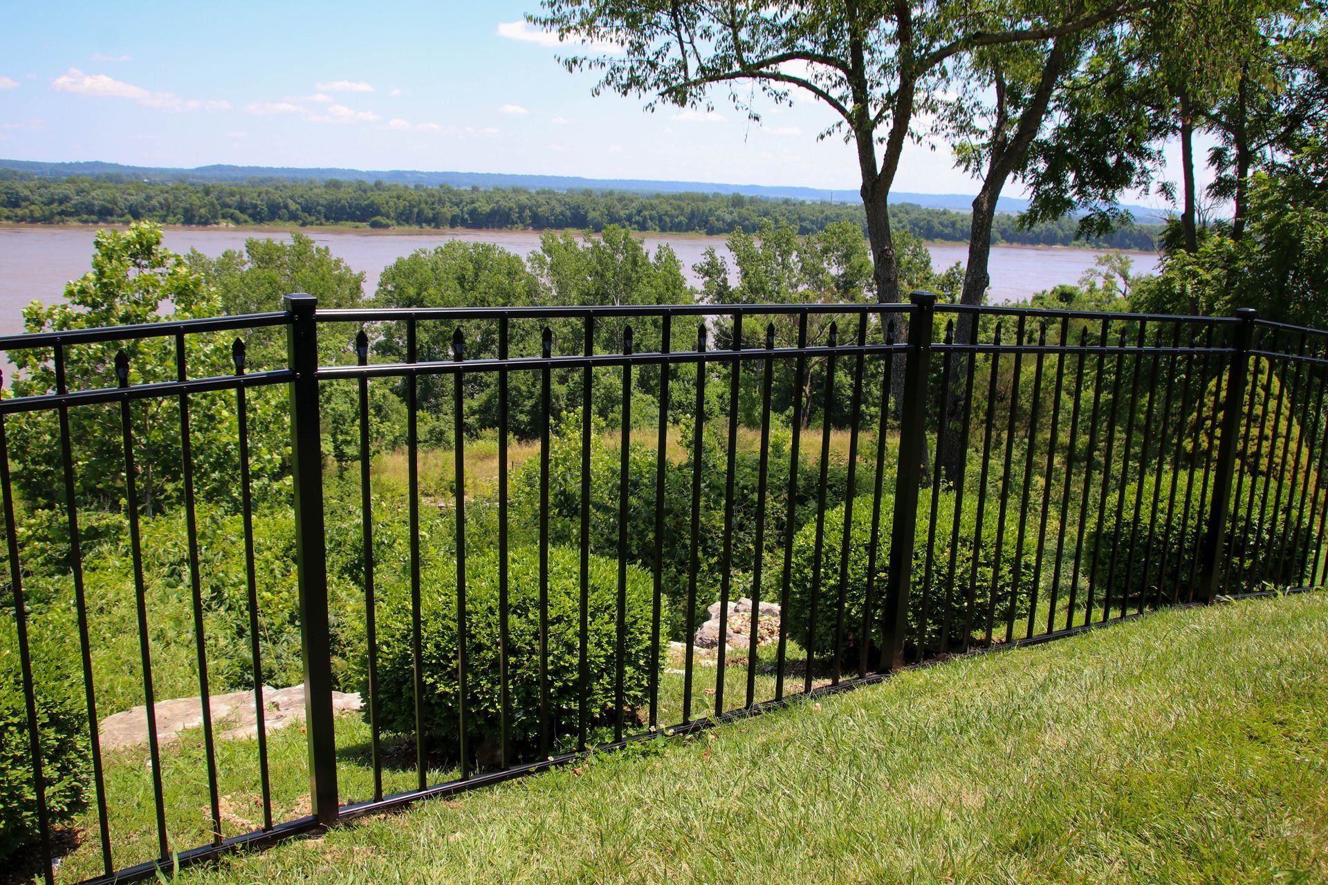 Black metal fence overlooking a river and treeline, sunny day.