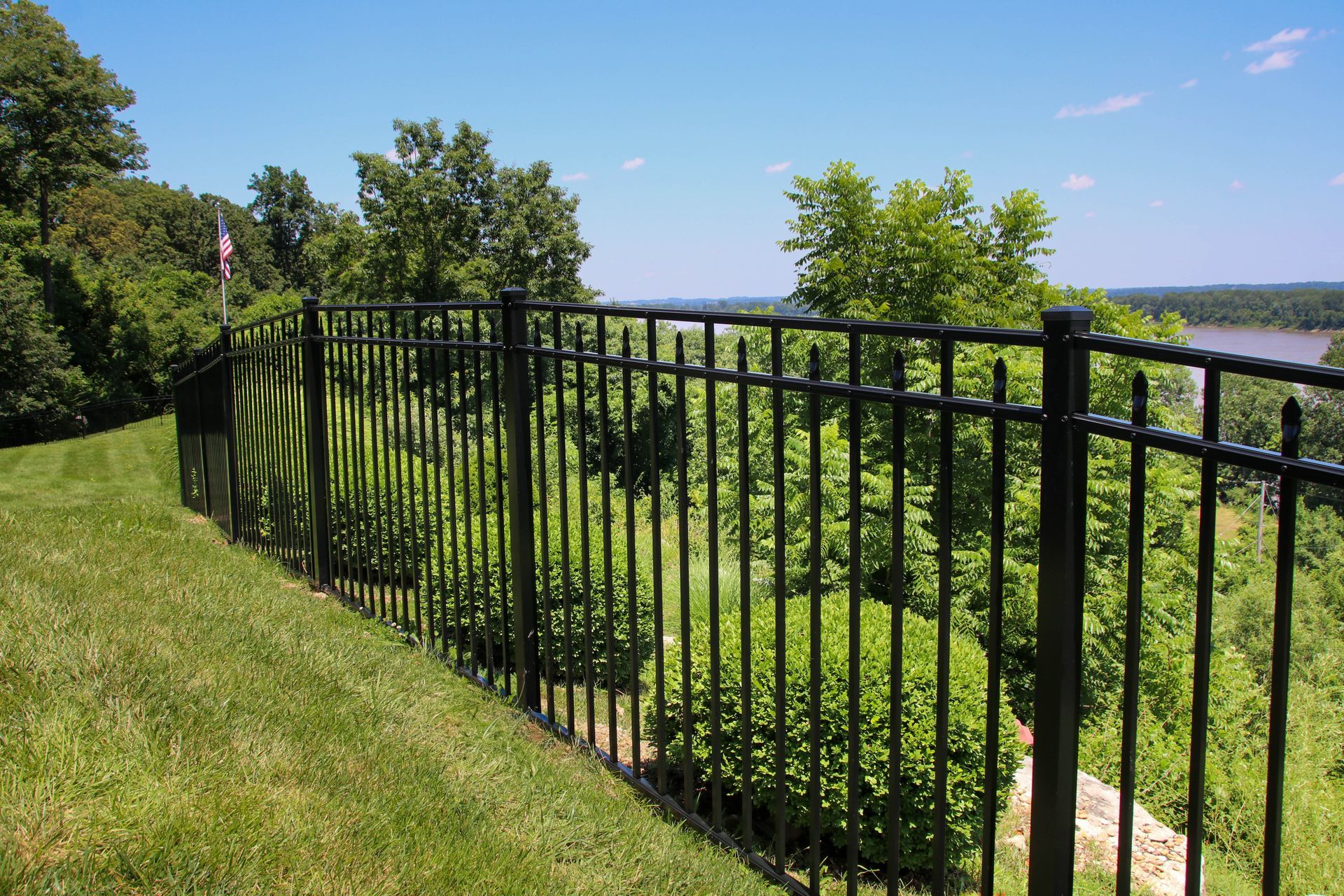 Black metal fence overlooking a green hillside and a river under a blue sky.