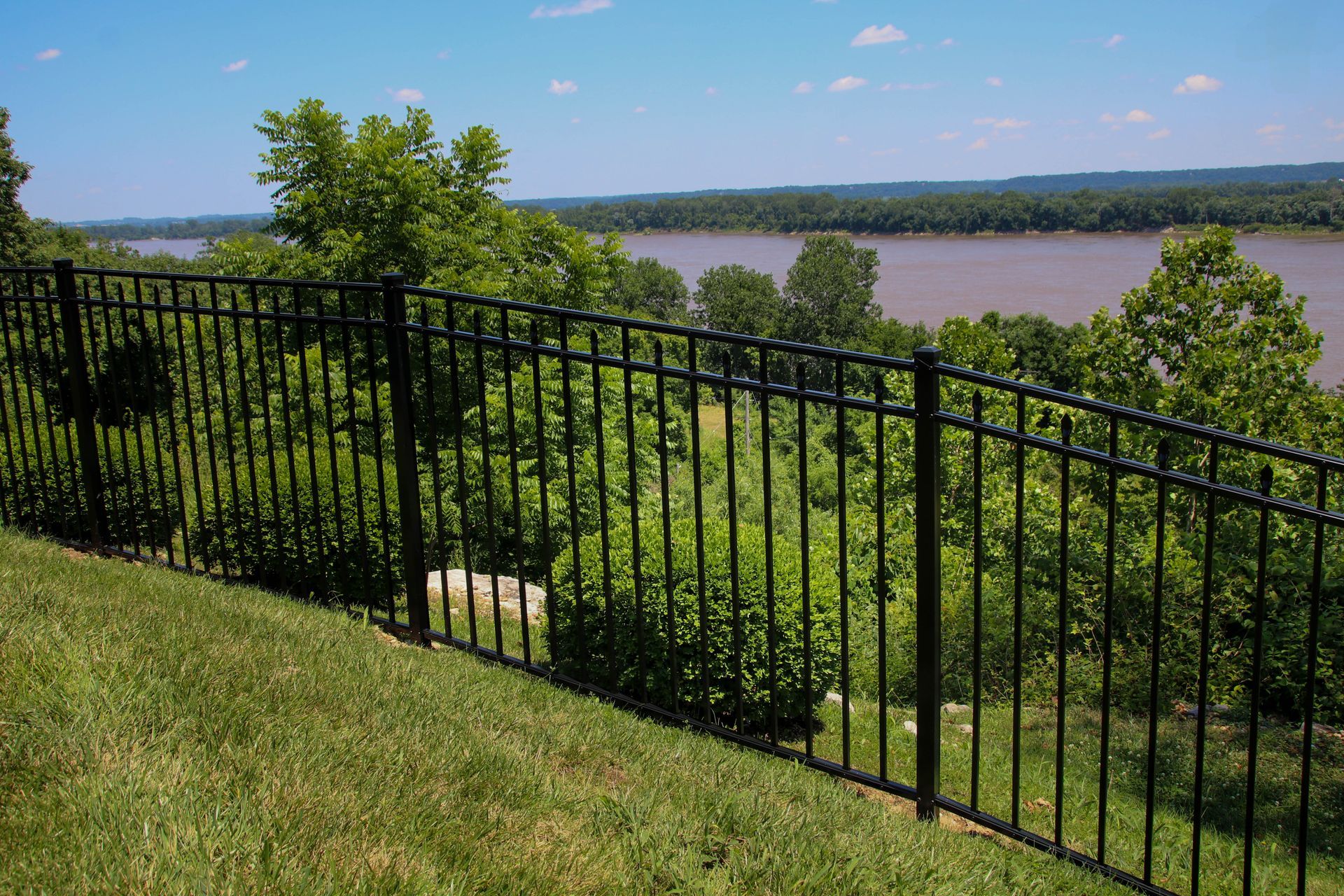 Black metal fence on a grassy hillside overlooking a river and treeline under a blue sky.