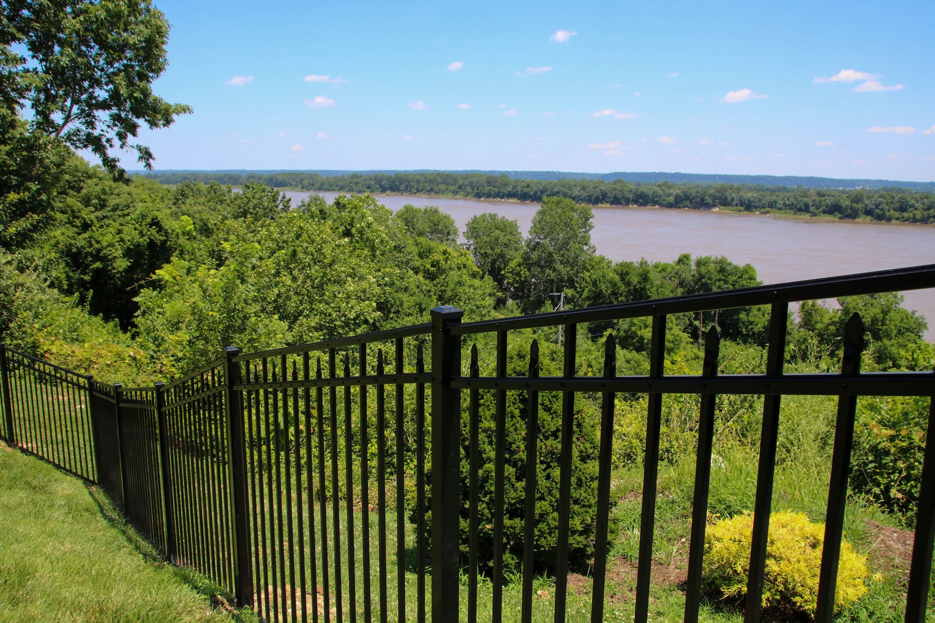 Black metal fence overlooking a wide river and green treeline under a blue sky.