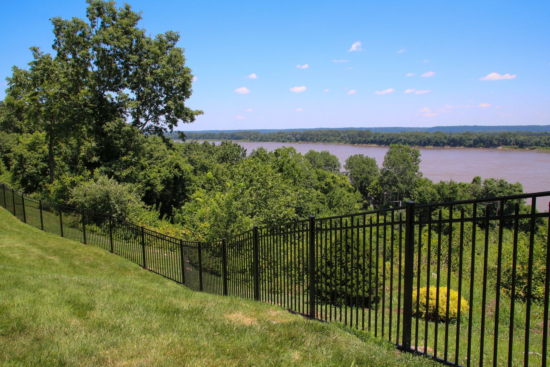 Black fence overlooking a river with green trees under a blue sky.