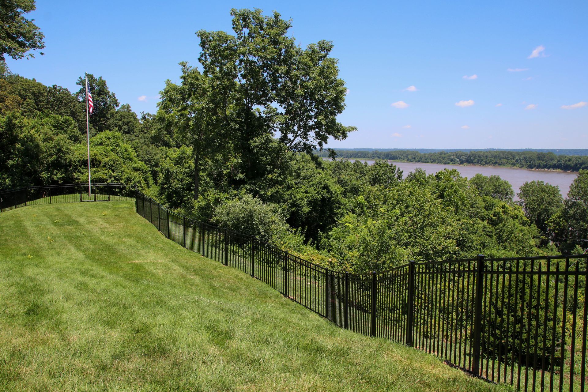 Green lawn slopes to black fence, trees, and river view under a blue sky.
