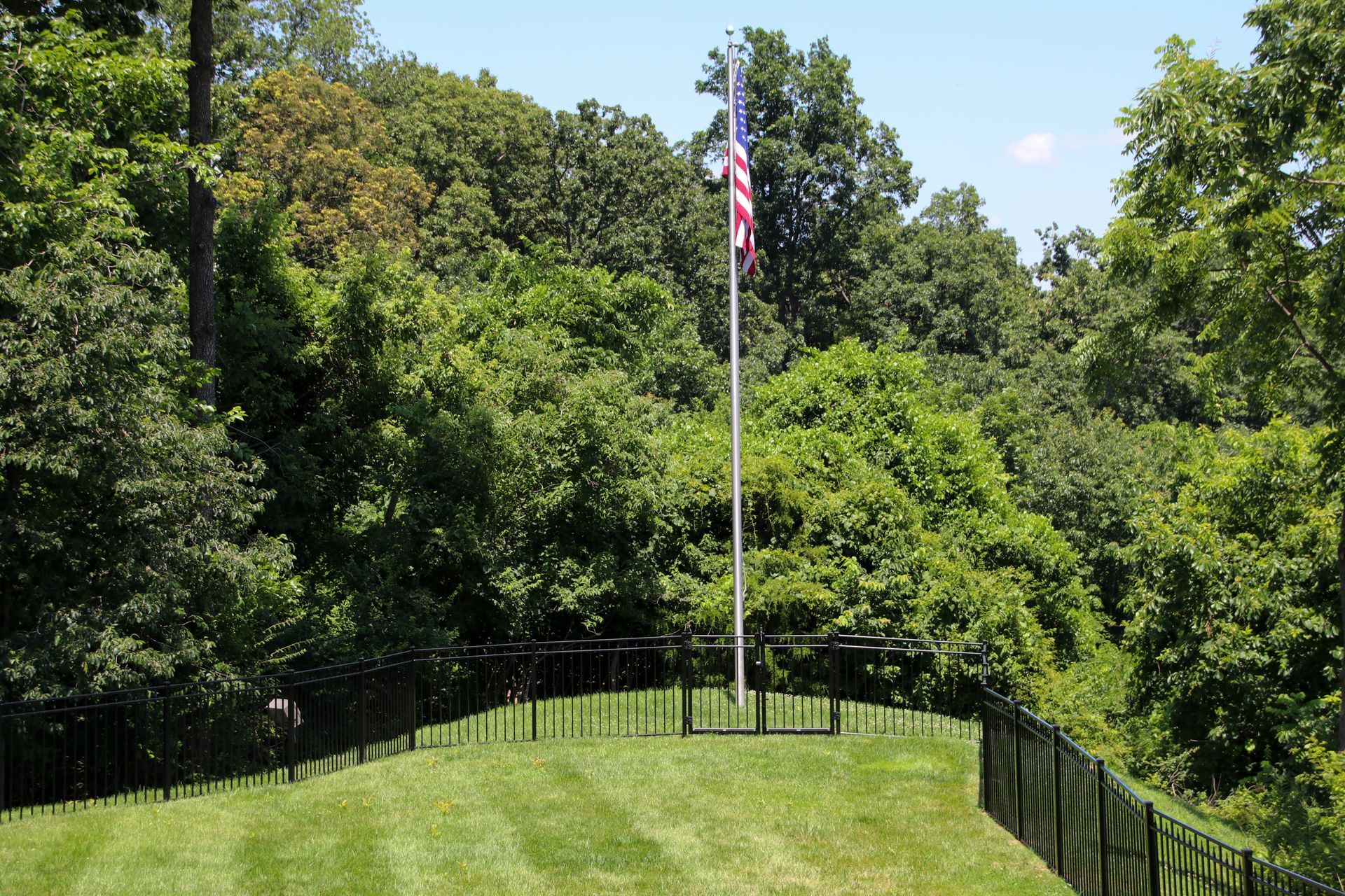 American flag on a tall pole, surrounded by green trees and a black fence, blue sky.
