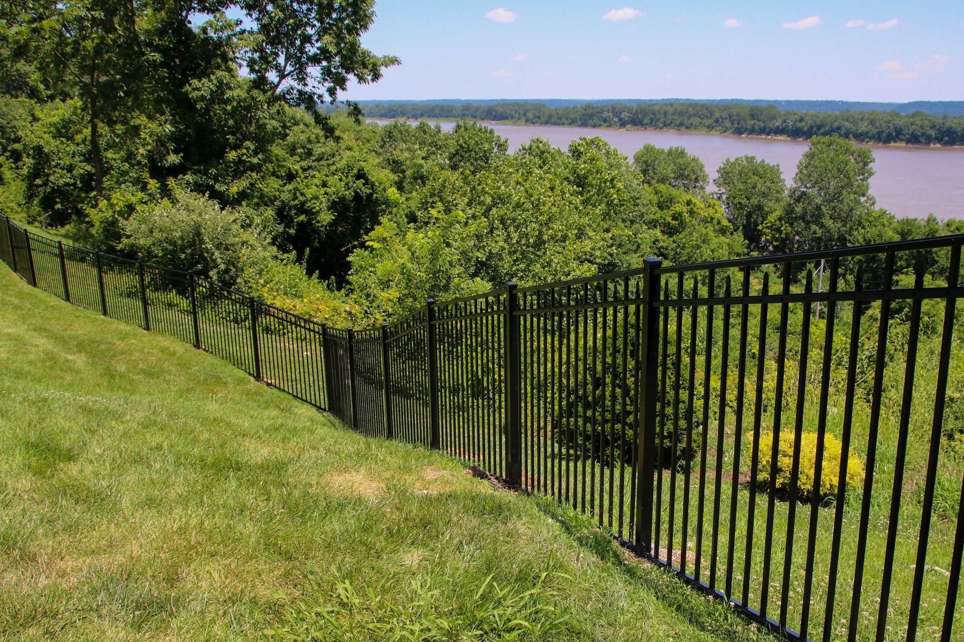 Black metal fence on a grassy hill overlooking a river and treeline on a sunny day.