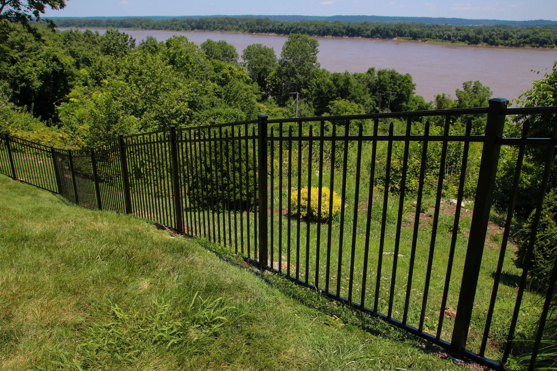 Black metal fence on a grassy hill overlooking a river and treeline.