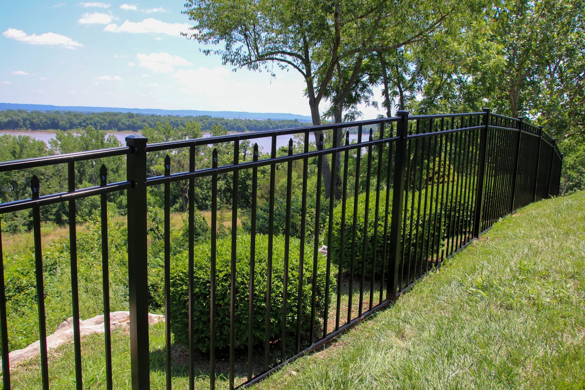 Black metal fence on grassy hill overlooking river and trees.