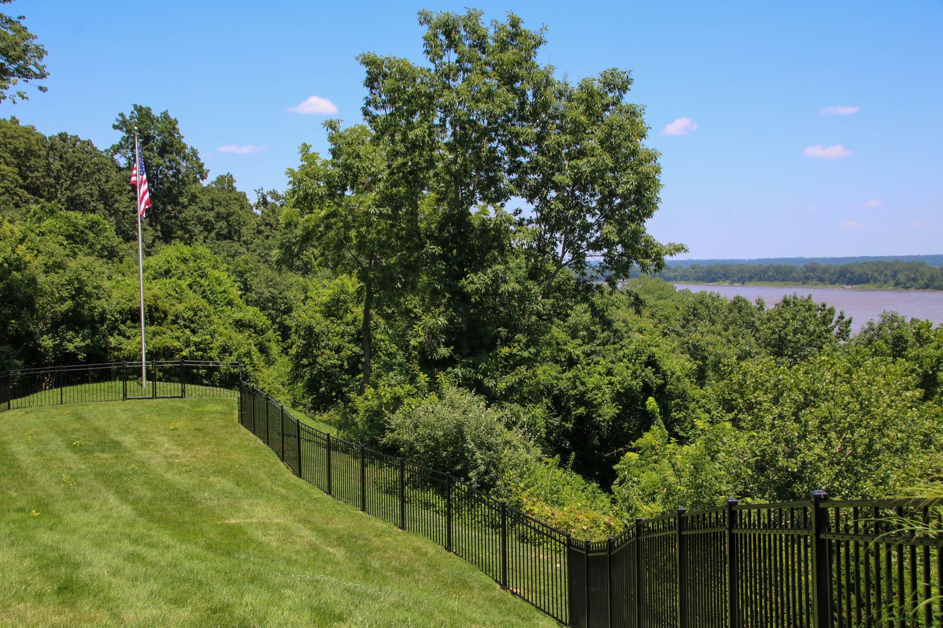 Lush green hillside with a black fence, American flag, and river view under a blue sky.