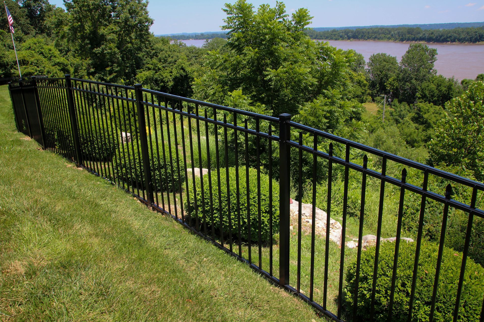 Black metal fence on a grassy hill overlooking a river and trees.