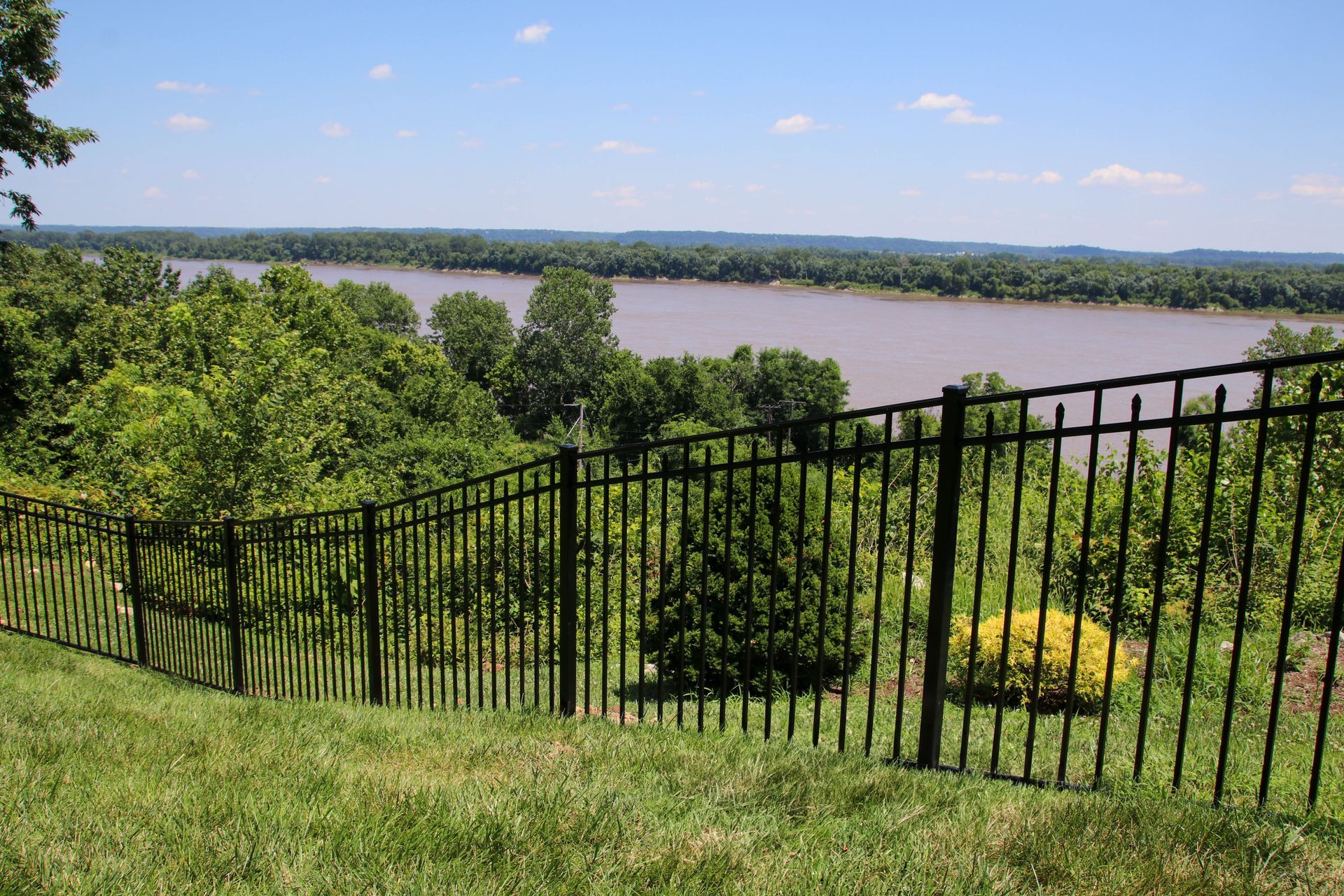 Black metal fence overlooks a wide, muddy river and green treeline under a blue sky.