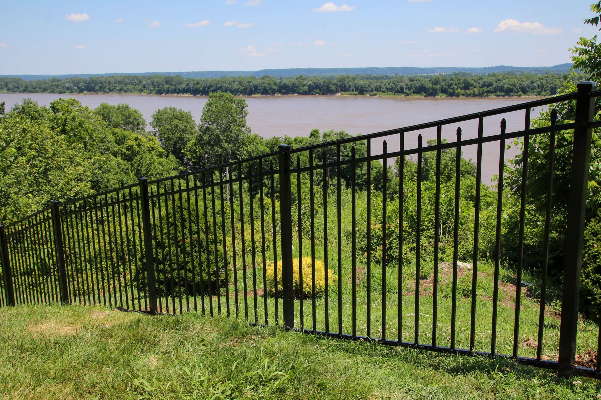 Black metal fence on a grassy hill overlooking a wide river and trees on a sunny day.