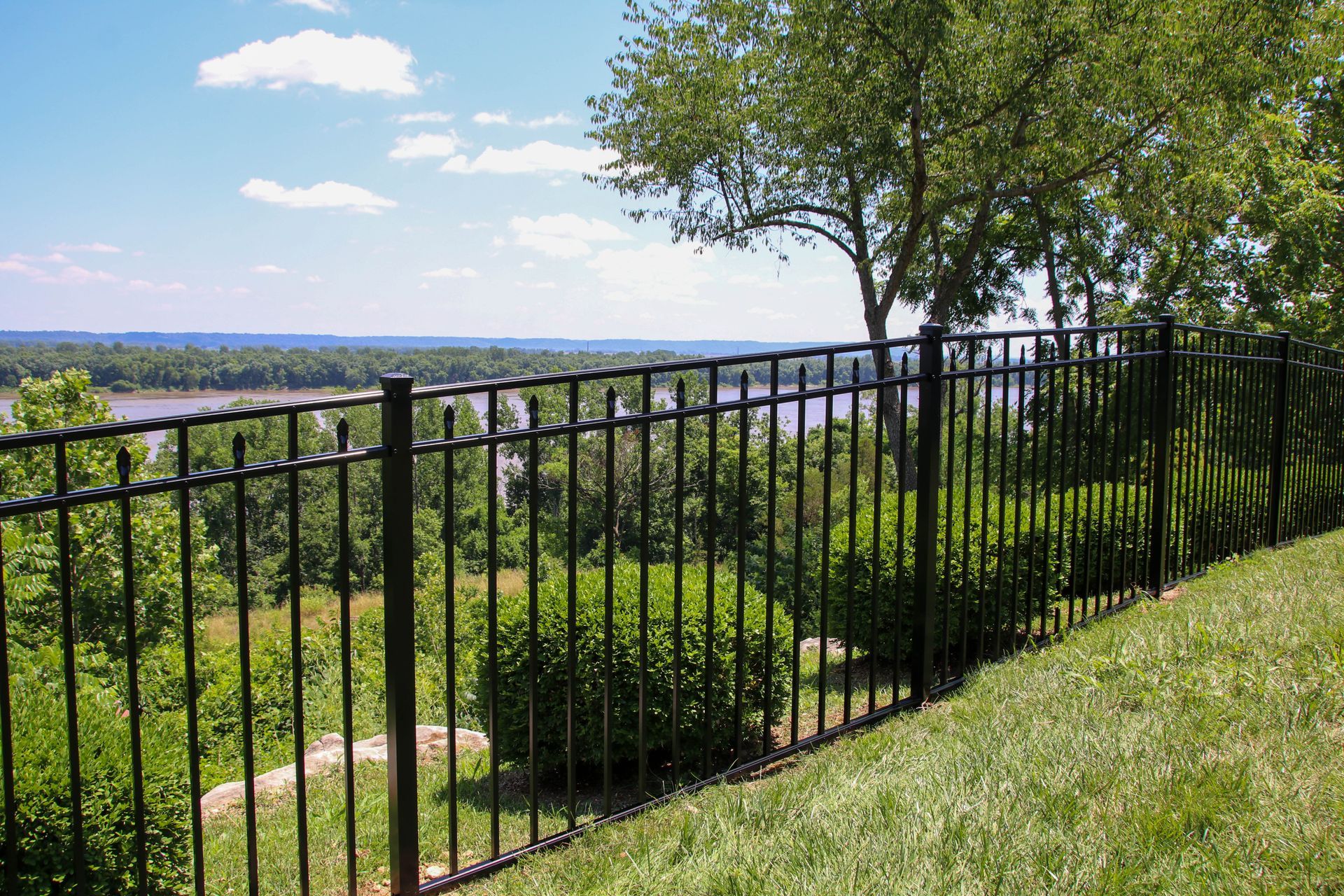 Black metal fence overlooks a river and trees on a sunny day.