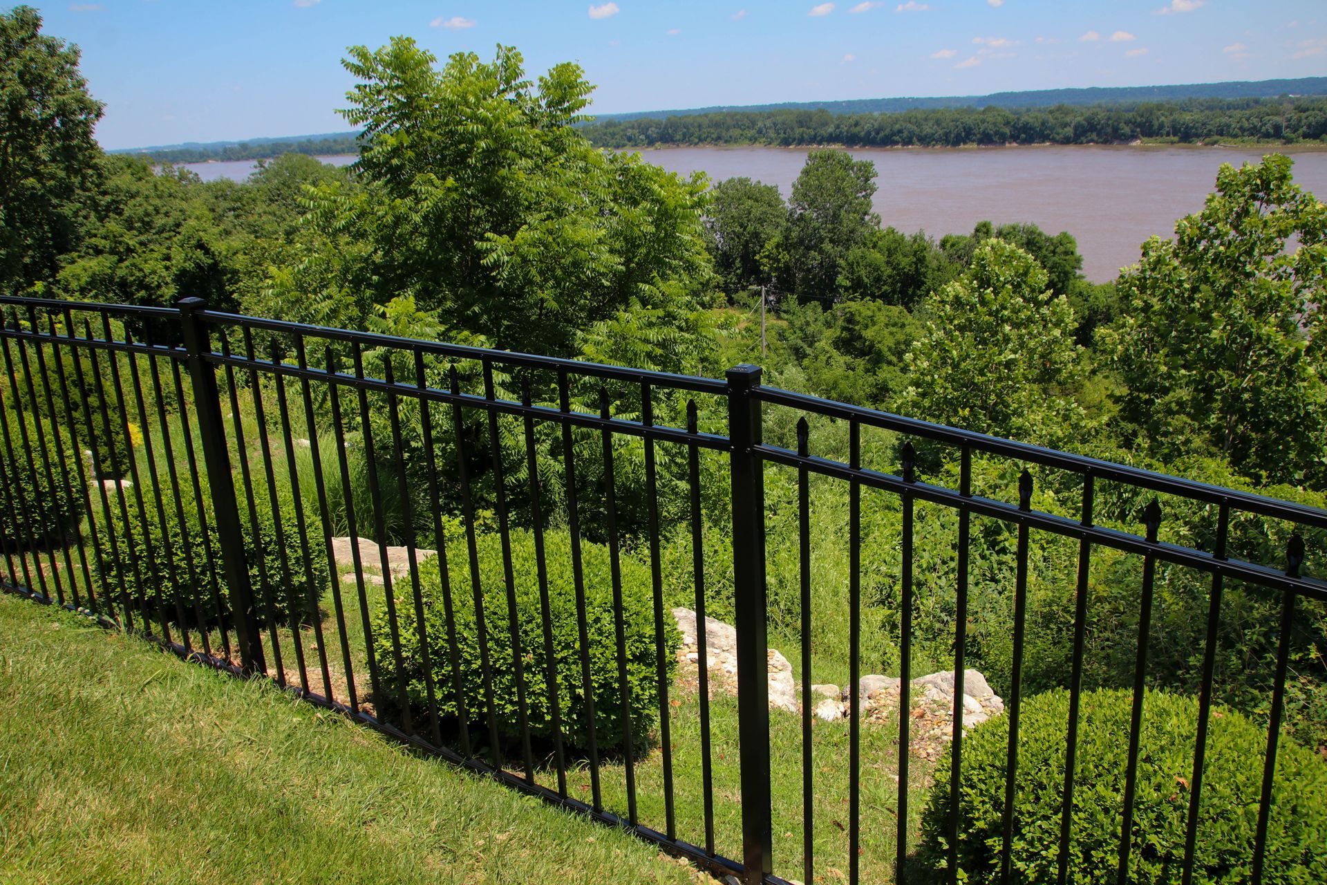 Black metal fence overlooking a river and green trees on a sunny day.