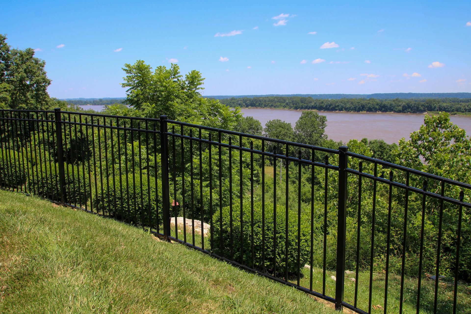 Black metal fence overlooking a river and treeline on a sunny day.