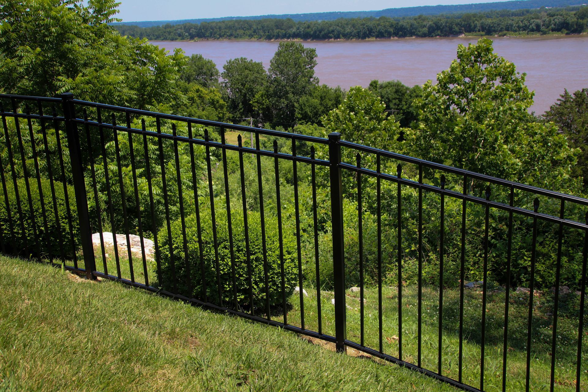 Black metal fence on a grassy hillside overlooking a river and trees on a sunny day.