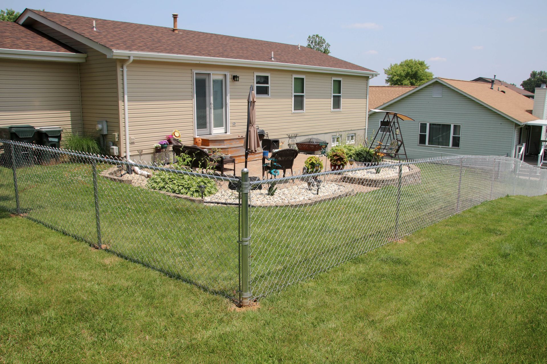 Chain-link fence in a backyard, with a house, flowers, and a patio visible in the background on a sunny day.
