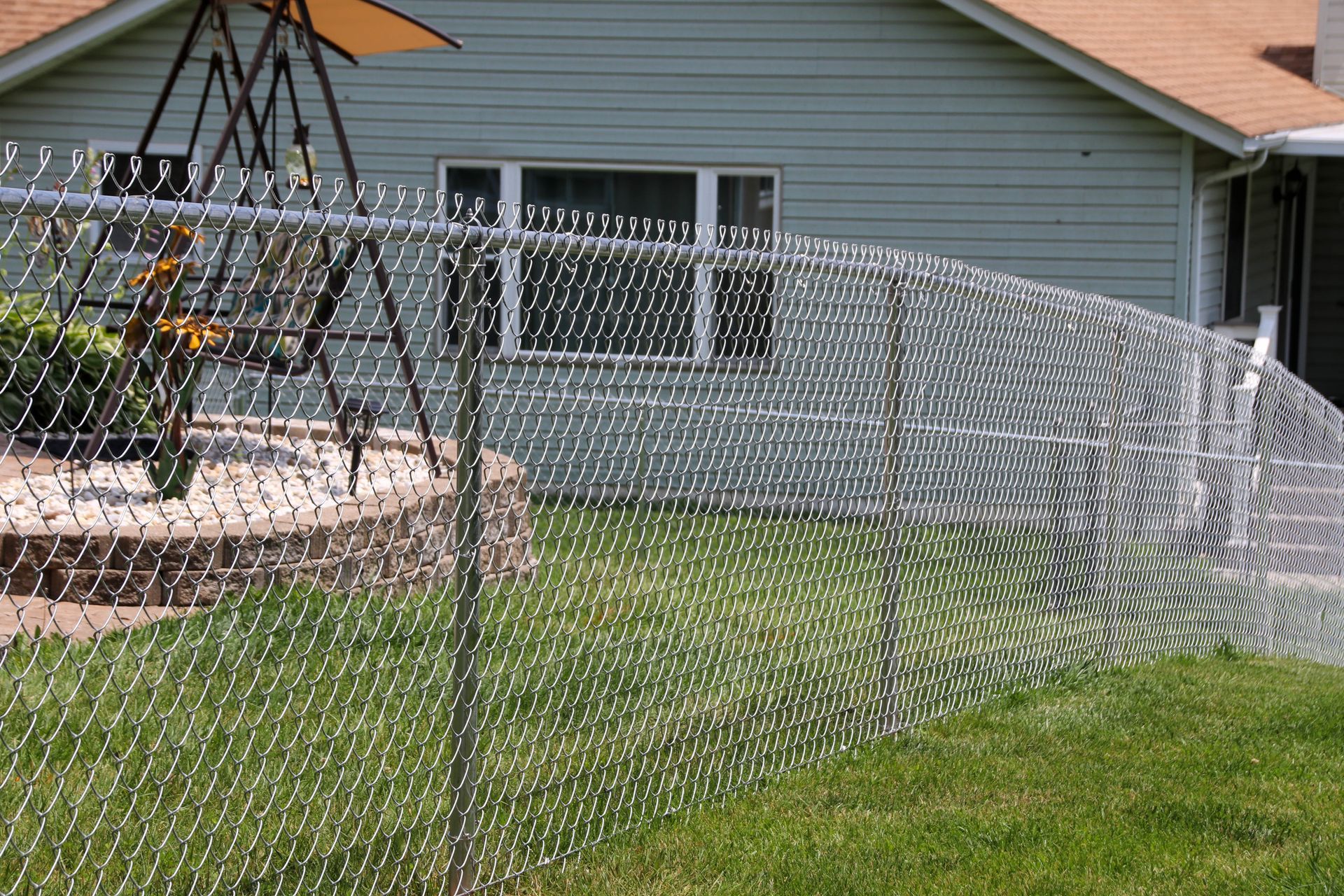 Chain-link fence in a yard curves toward a light green house with brown roof.