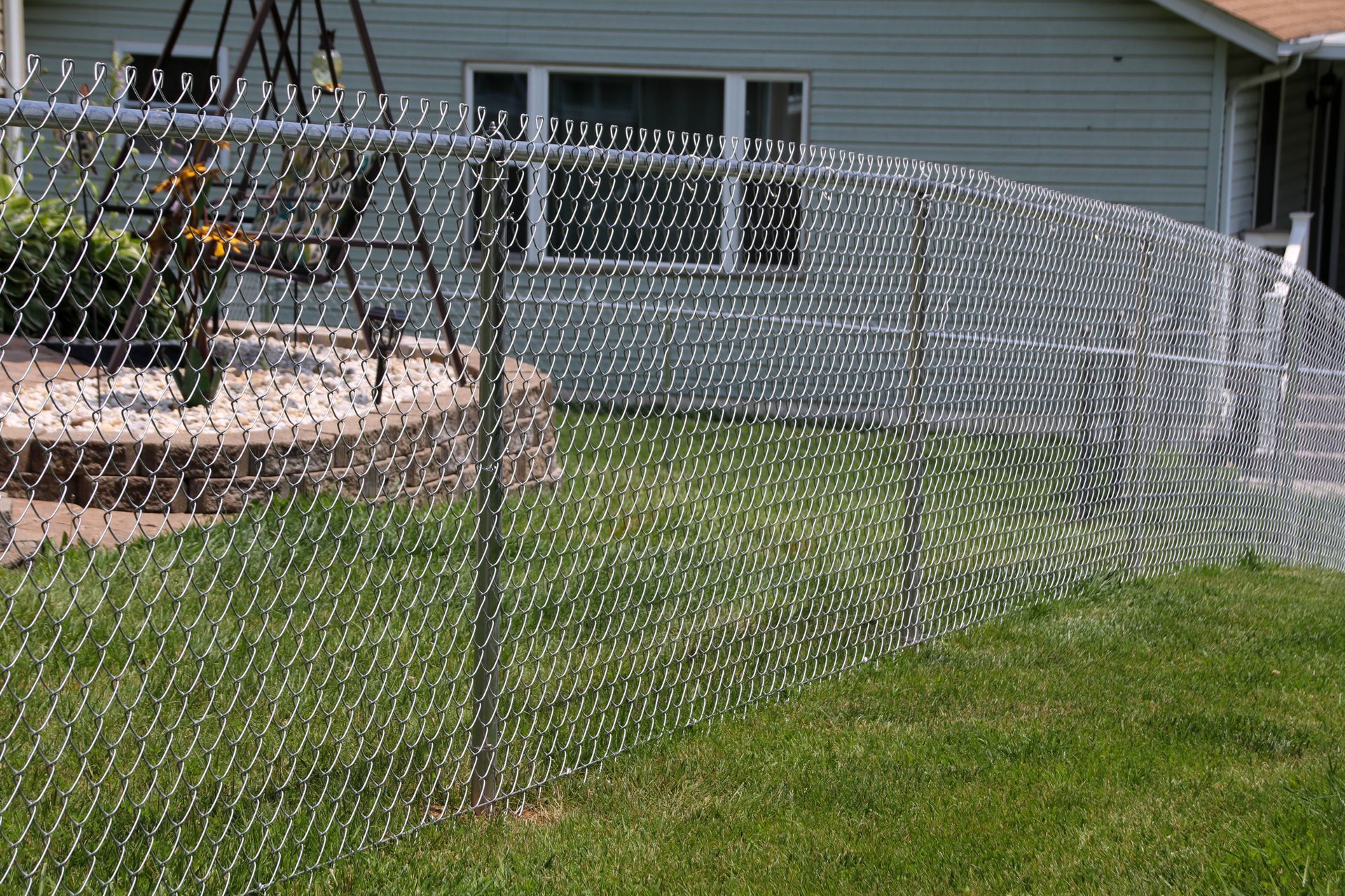 Chain-link fence in front of a house, with green grass in the foreground and a swing set visible.