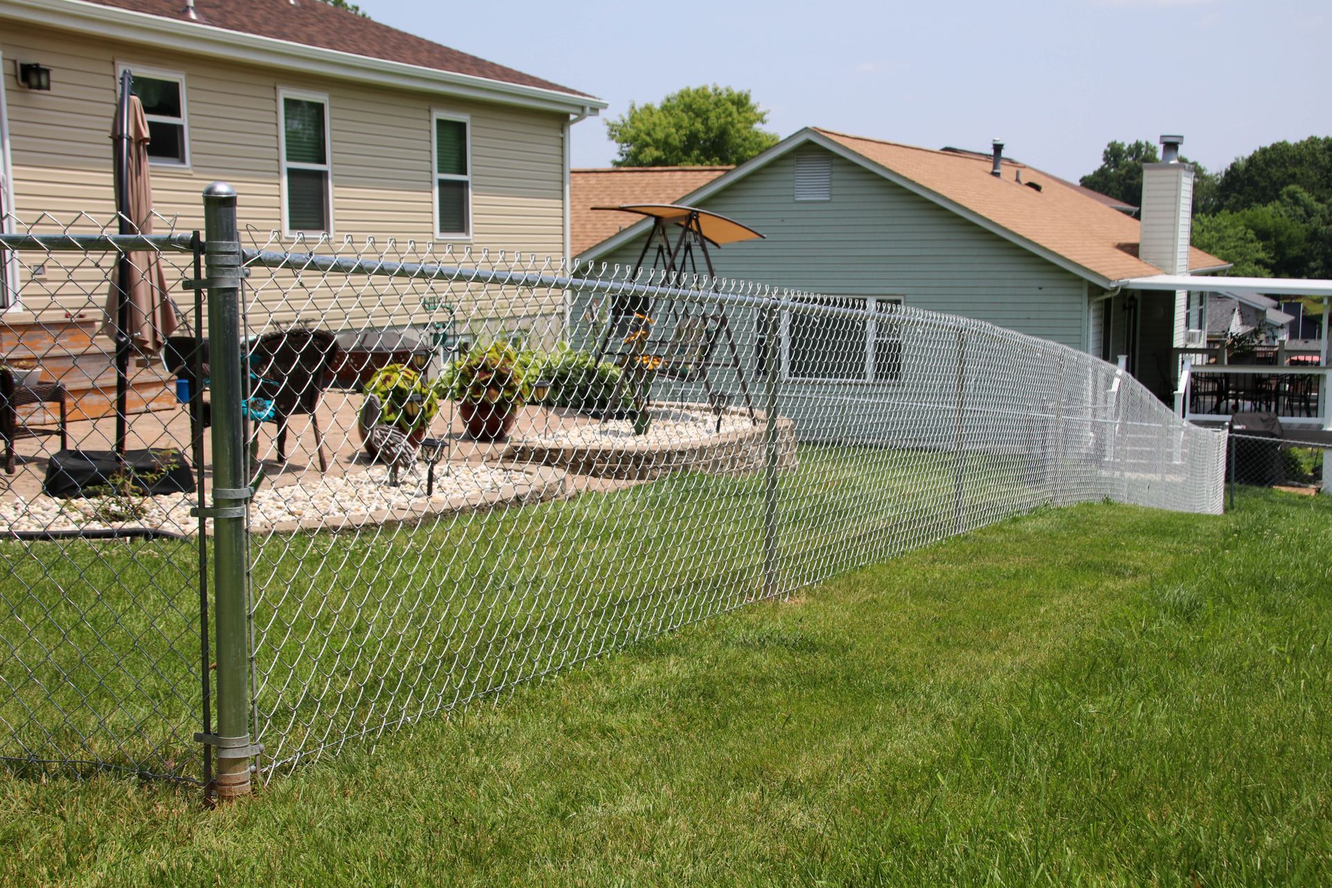 Chain-link fence in a grassy backyard, curving between houses under a bright sky.