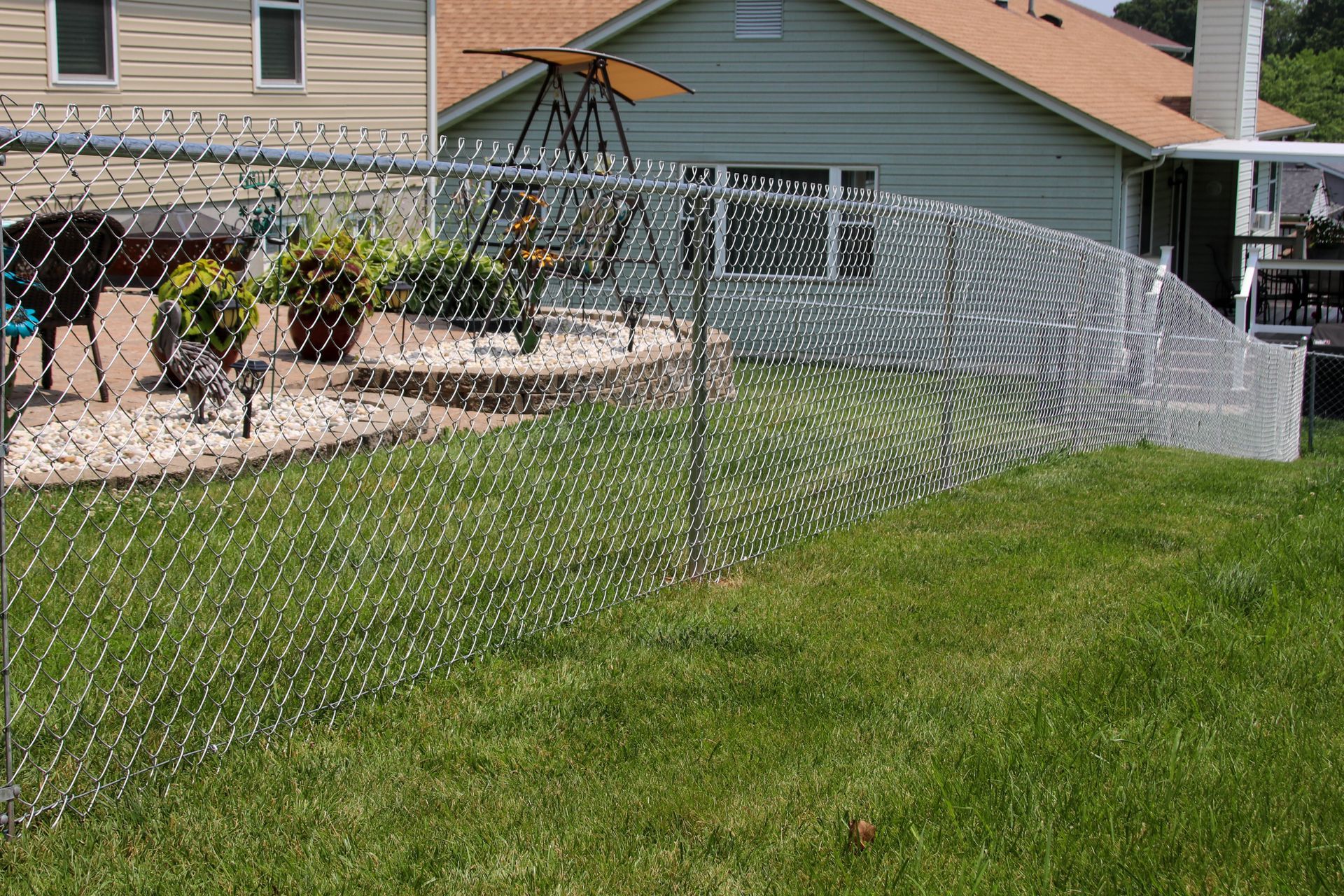 Chain-link fence in a backyard; green grass, plants, and a swing set are visible.