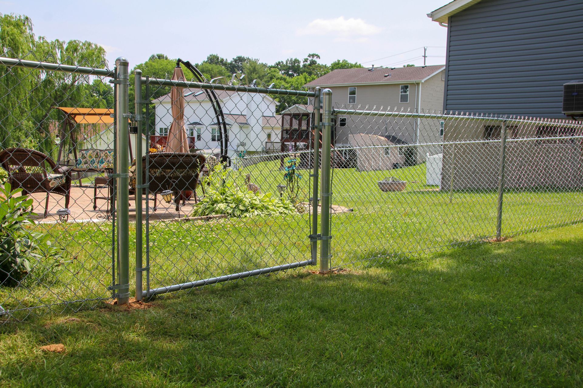 Chain link fence with open gate in a grassy backyard. Houses in background.