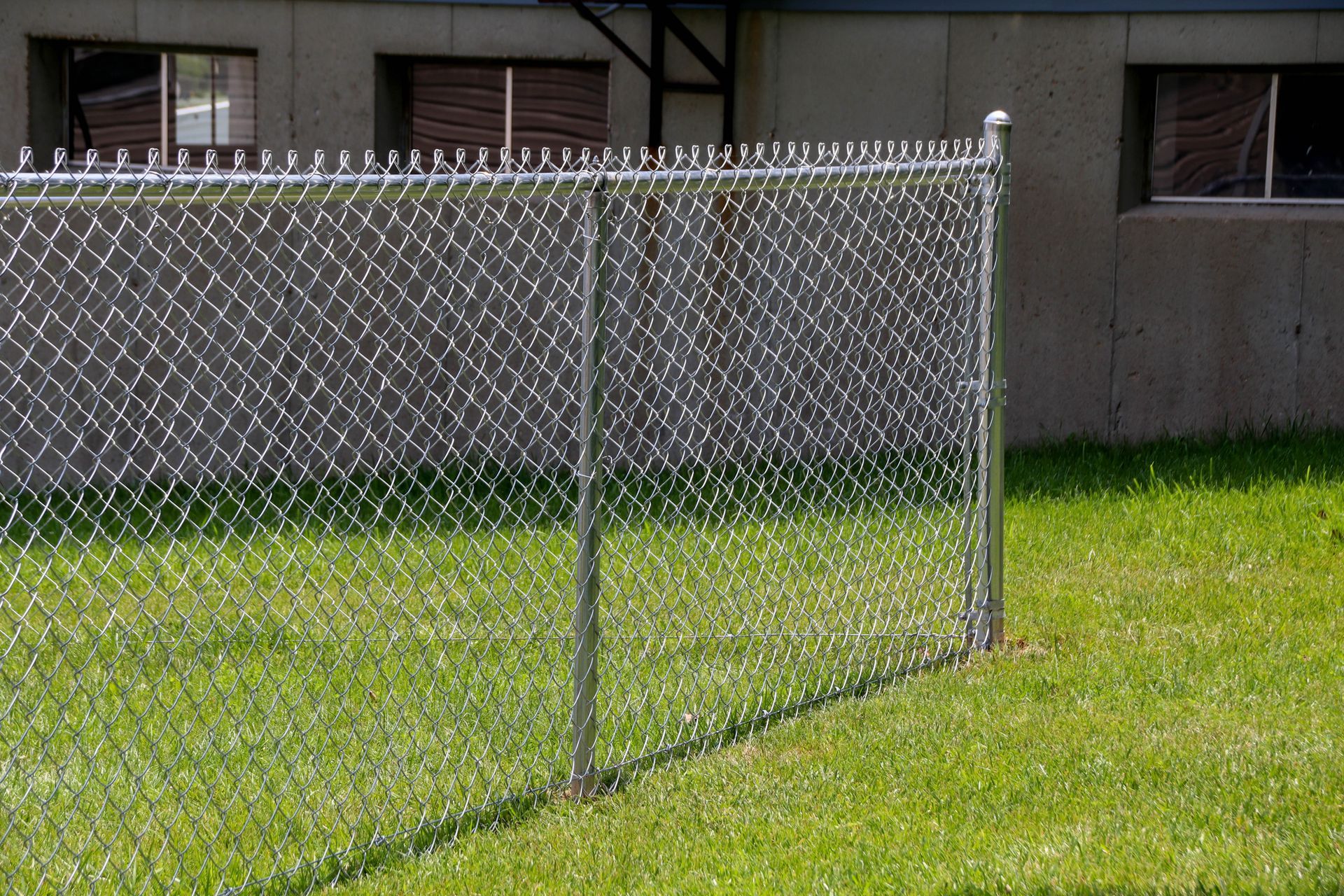 Chain-link fence with razor wire, set on green grass, next to a concrete building with windows.