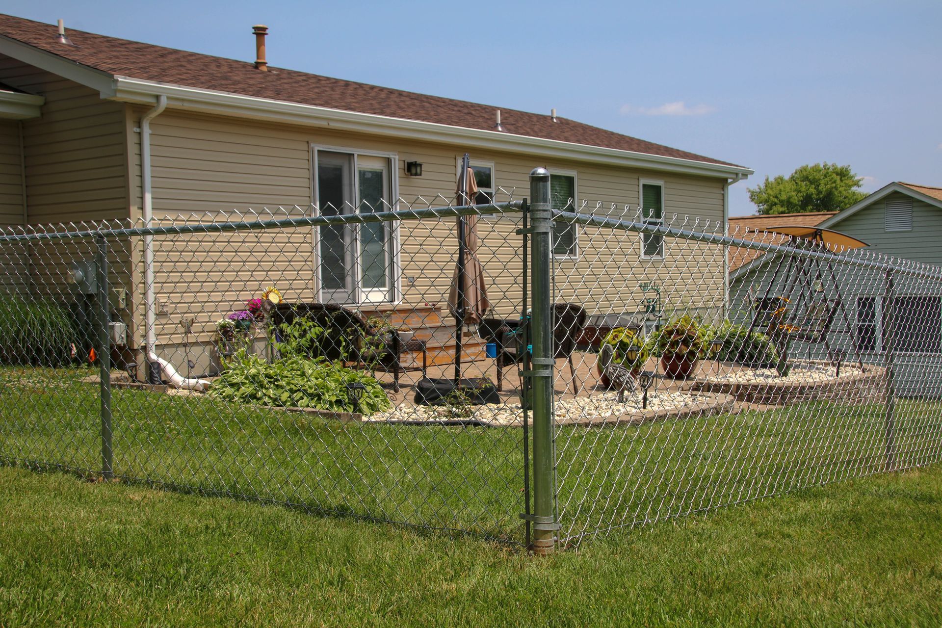 Chain-link fence in a backyard; beige house with a patio in the background, green grass and blue sky.