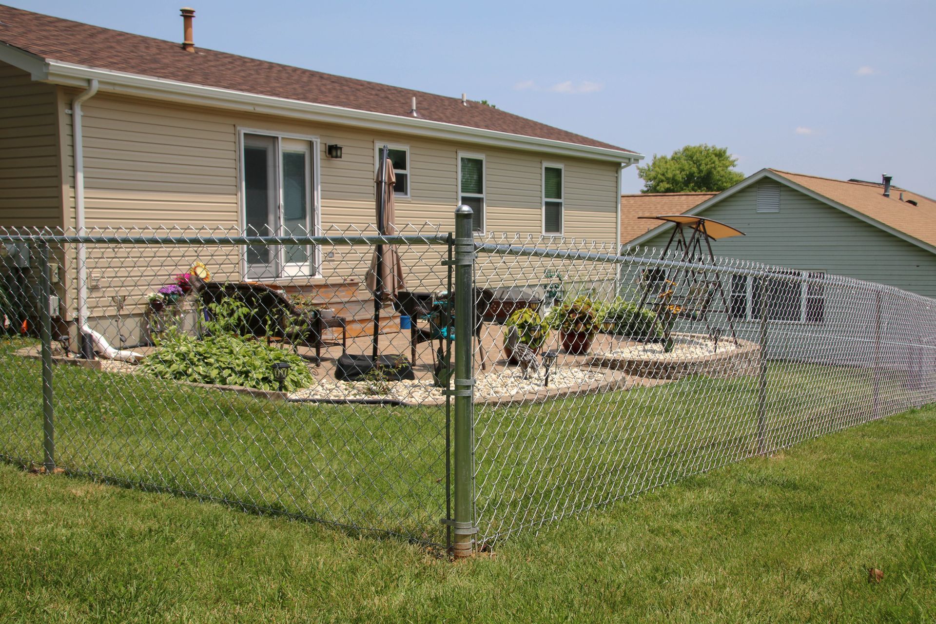 Chain-link fence surrounds a backyard with a house, deck, and plants. Sunny day, green grass.