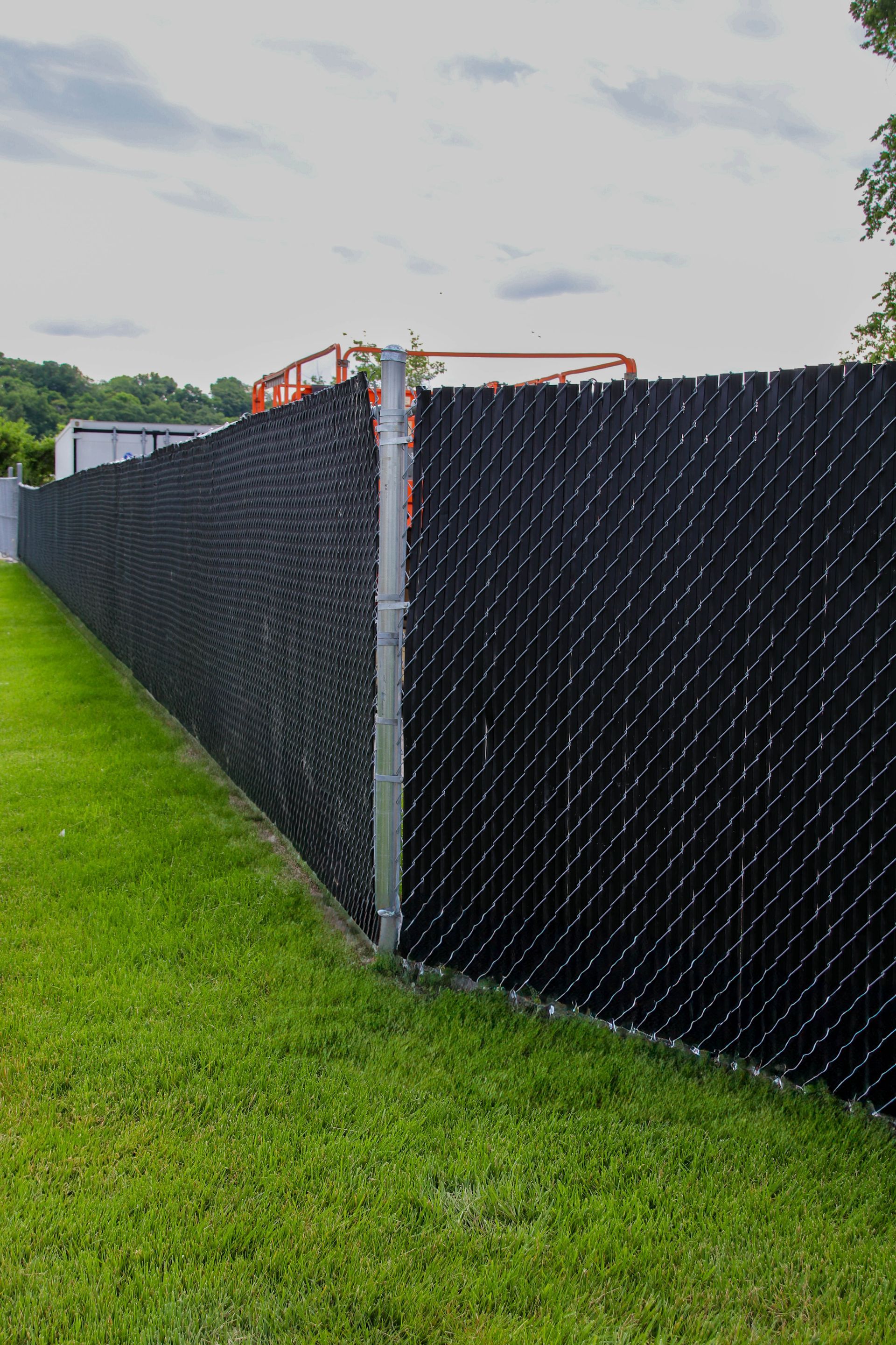 Black privacy fence with green grass in the foreground under a cloudy sky.