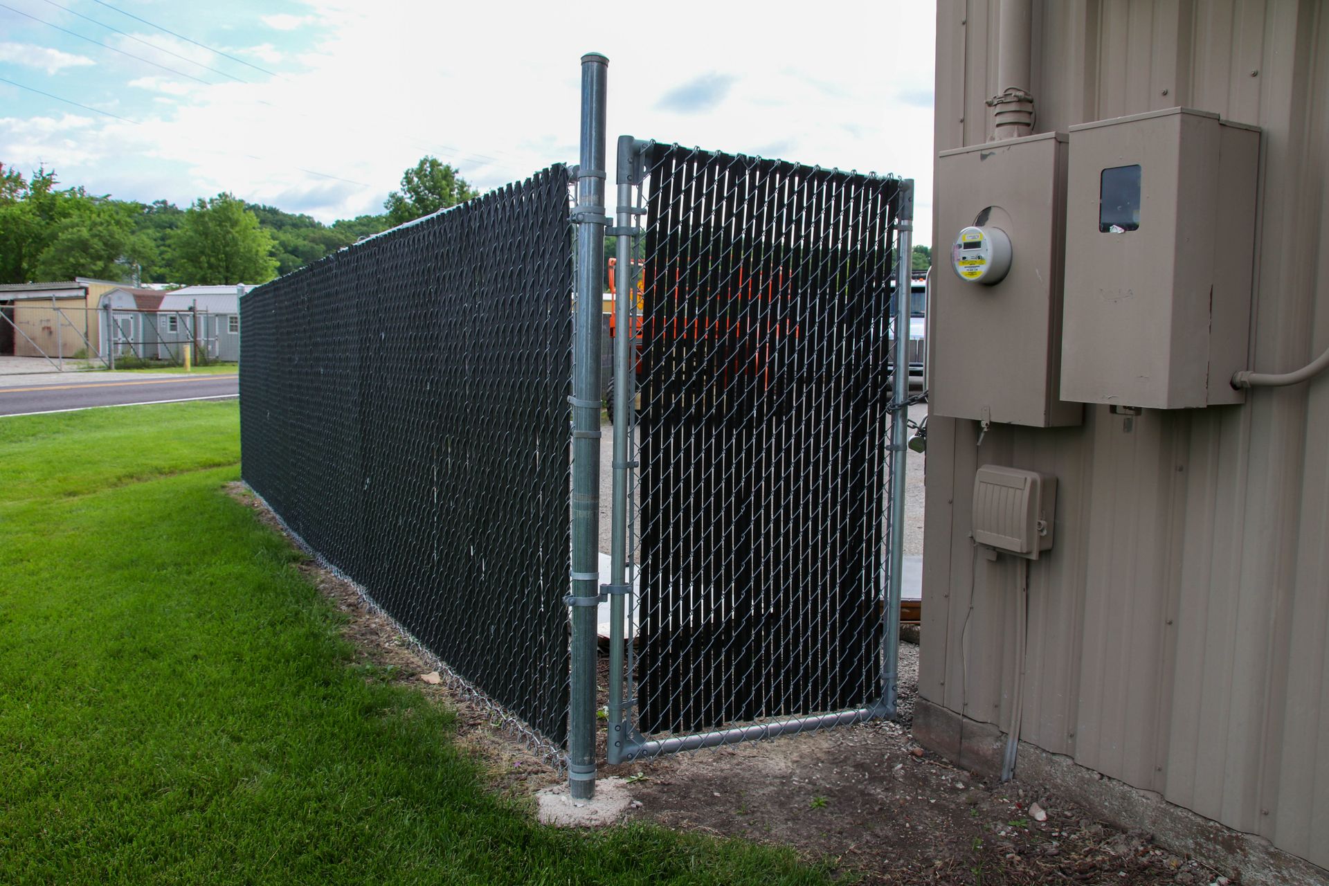 Black chain-link fence with privacy slats, attached to a building with electrical boxes, beside a grassy area.