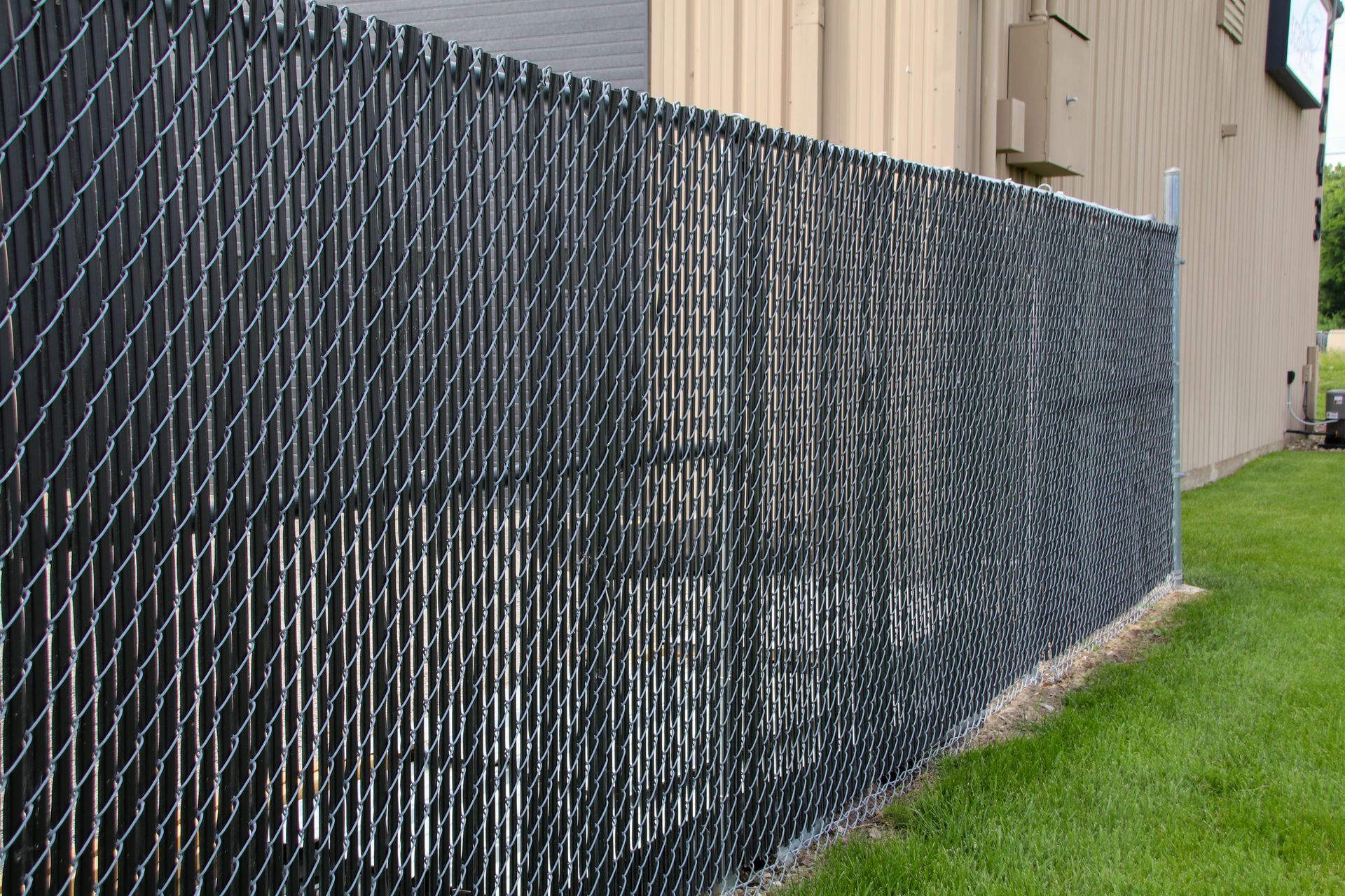 Black privacy slats inserted in a chain-link fence, beside a building and green lawn.