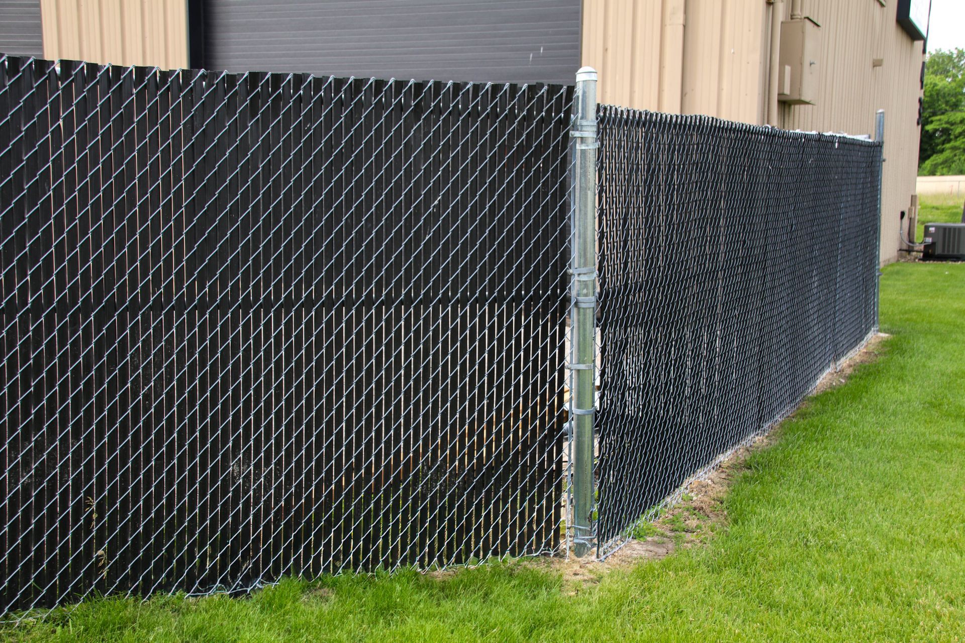Black chain-link fence with privacy slats, bordering a grassy area next to a tan building.