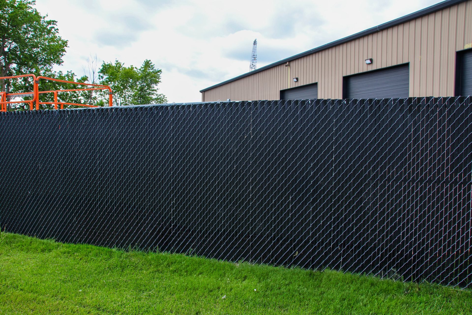 Black chain-link fence with privacy slats in front of a tan industrial building and green grass.