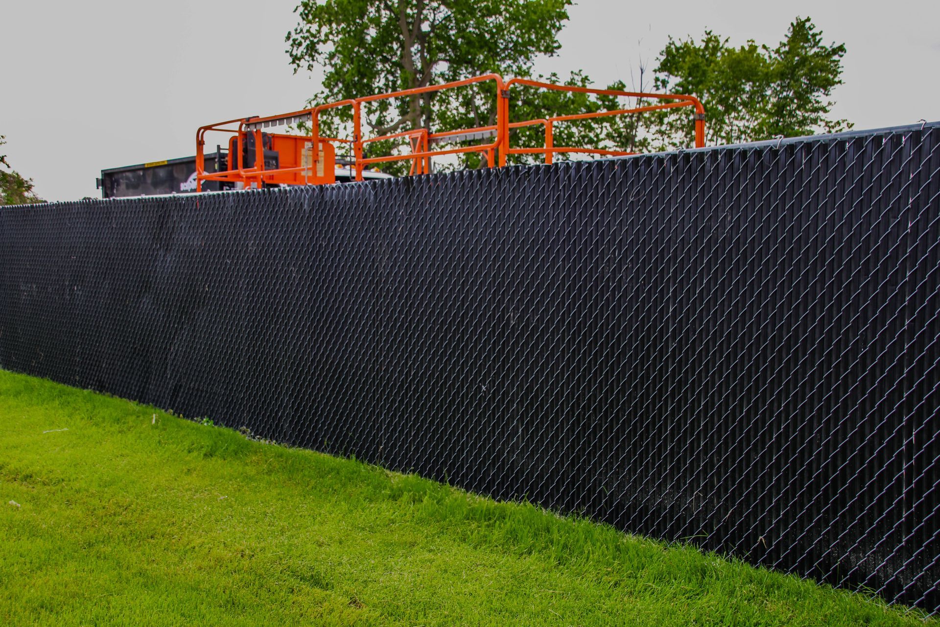 Black chain-link fence with privacy slats bordering a grassy area, a lift is visible in the background.