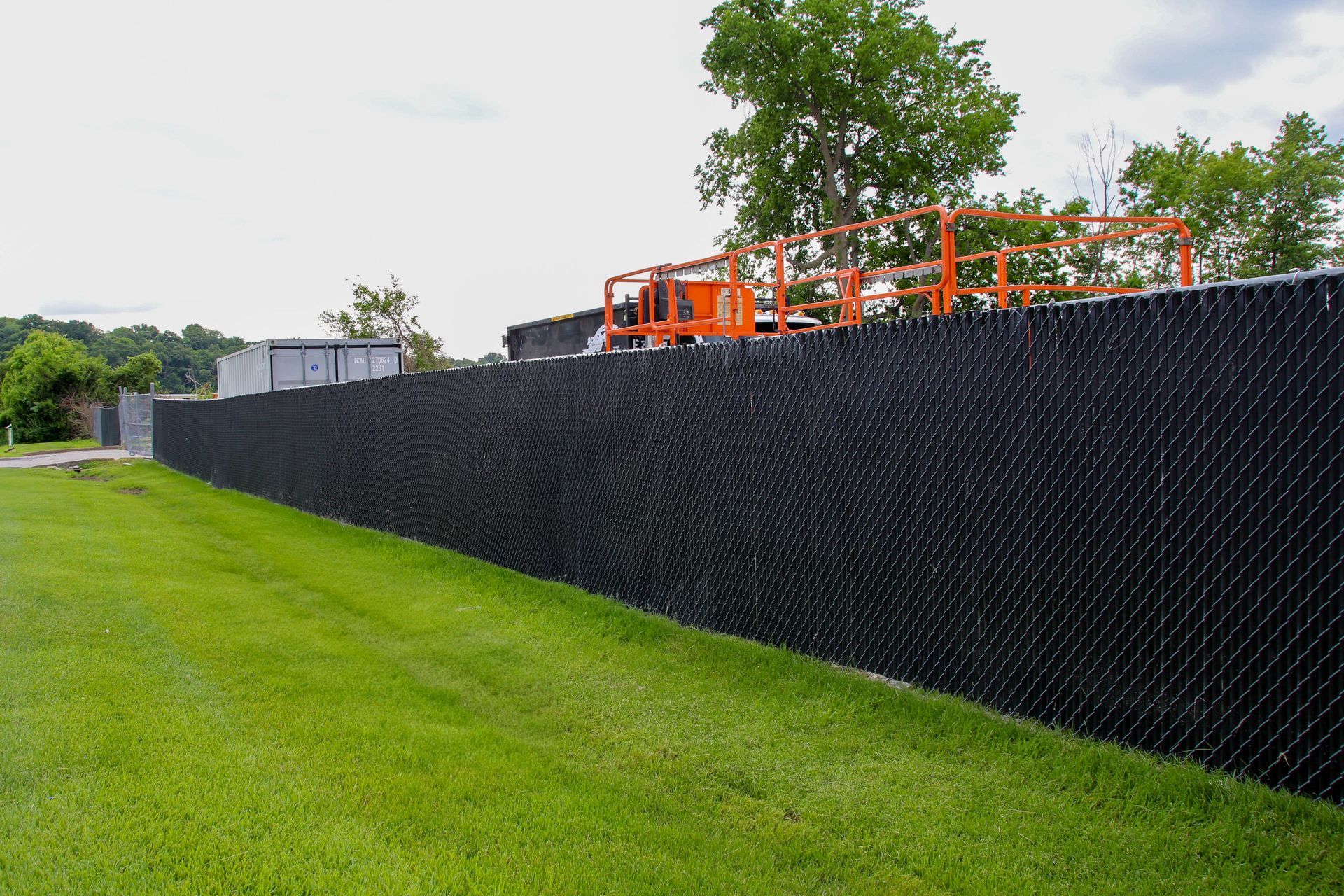 Black chain-link fence with privacy slats along green grass. An orange lift is visible over the fence.