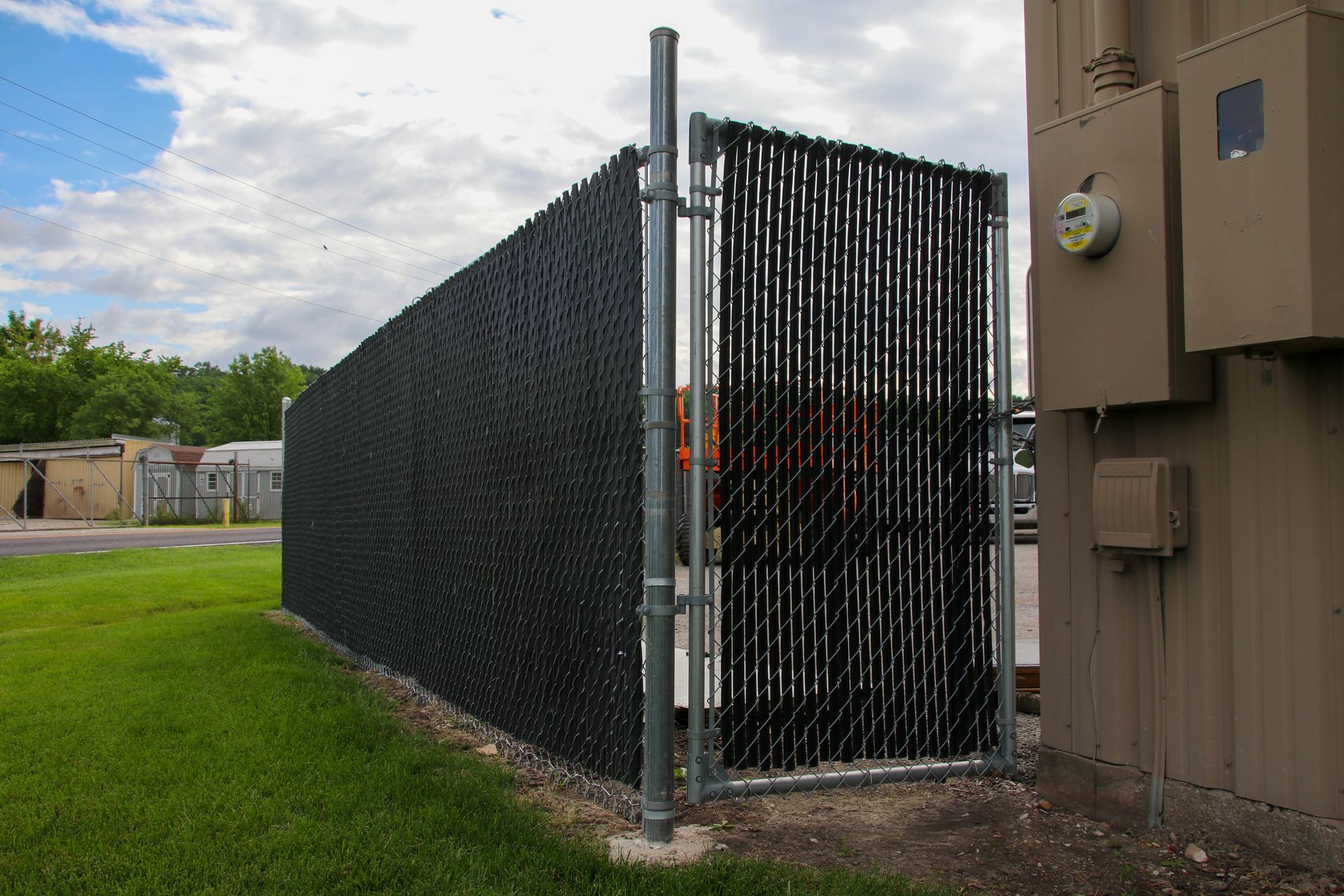 Black privacy fence around equipment, on green grass, next to a building on a cloudy day.