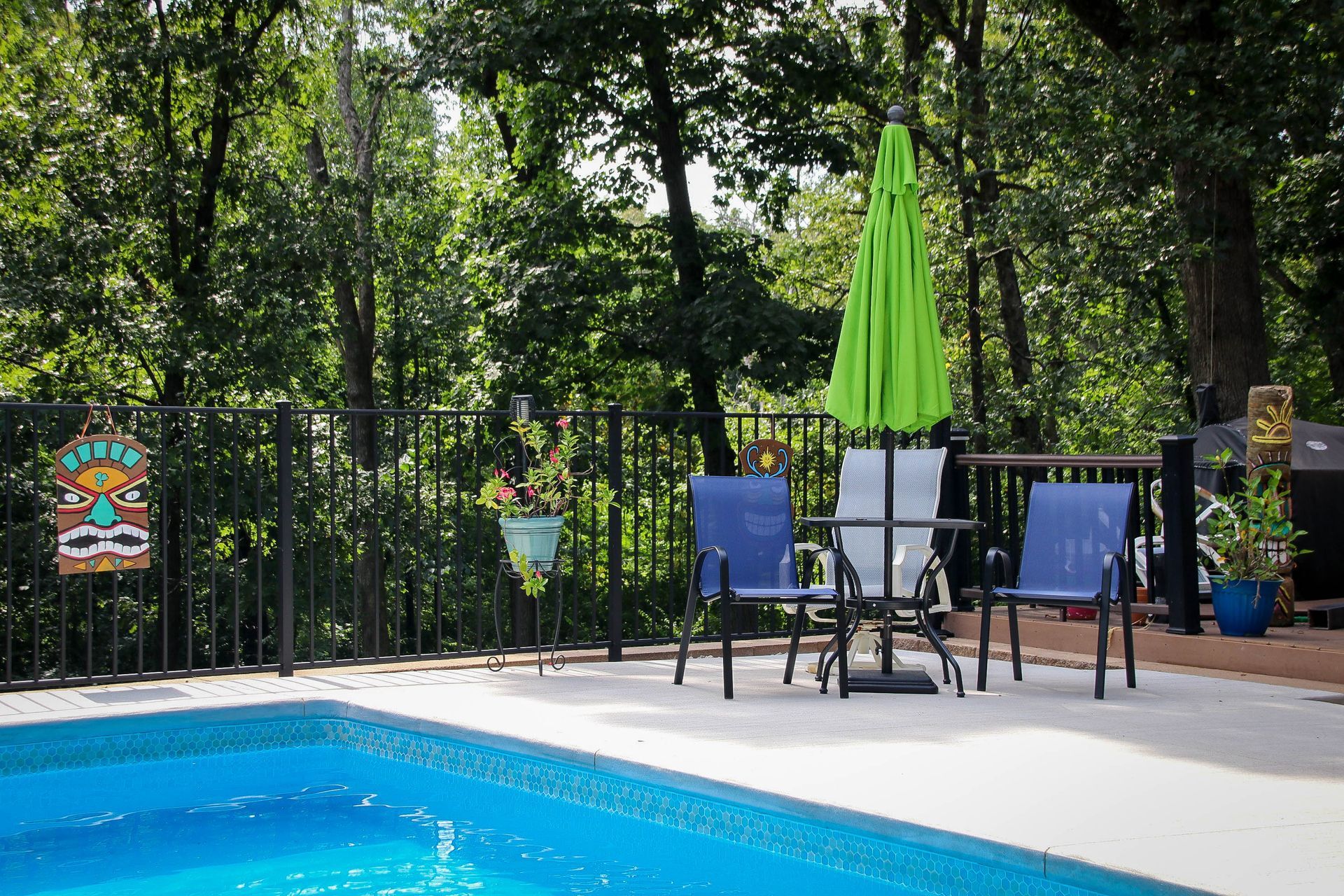 Poolside patio with blue chairs, a table, and green umbrella. Black fence and lush trees in the background.