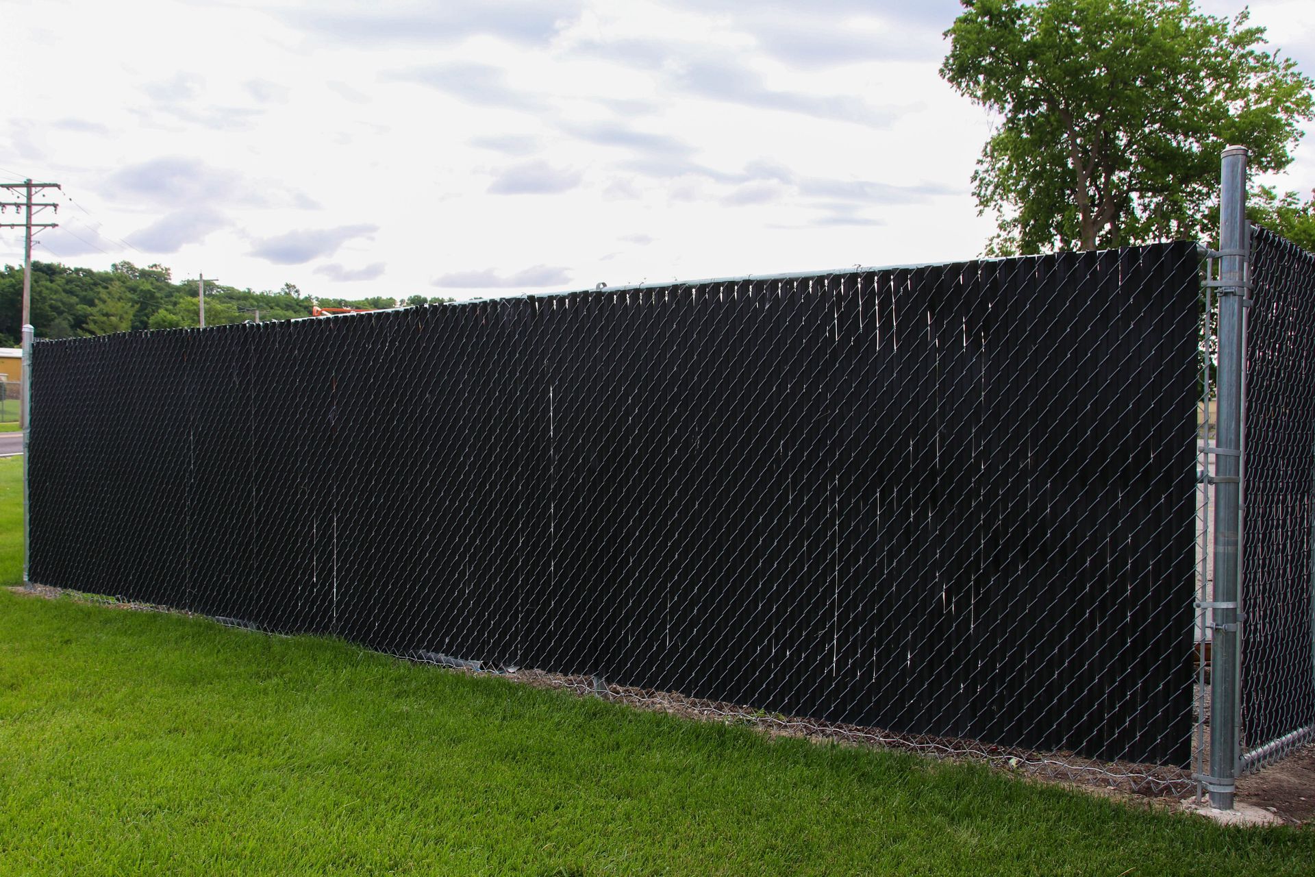 Black privacy slats inserted into a chain link fence, on green grass, under a cloudy sky.