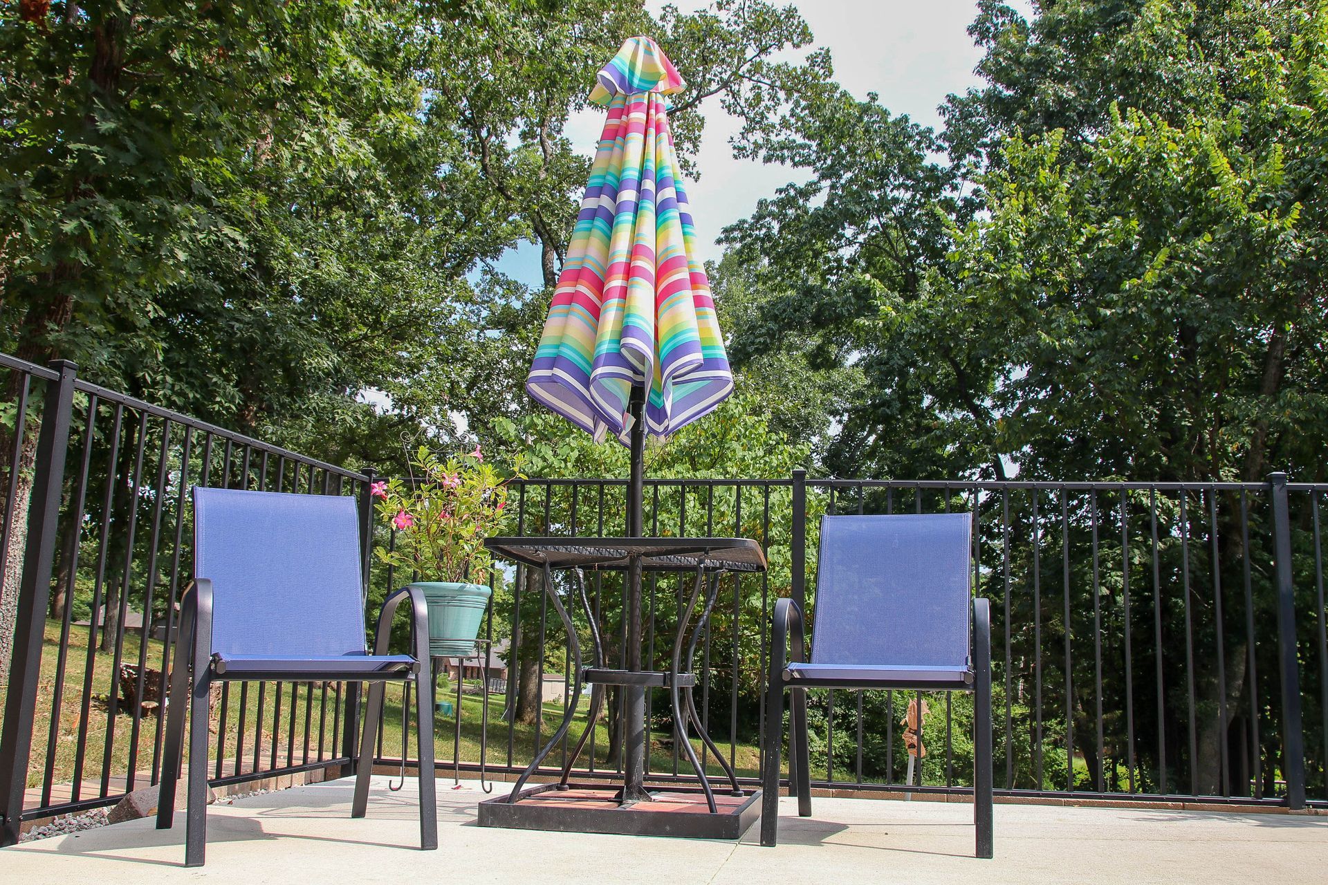 Patio with two blue chairs, small table, and colorful umbrella; surrounded by trees.