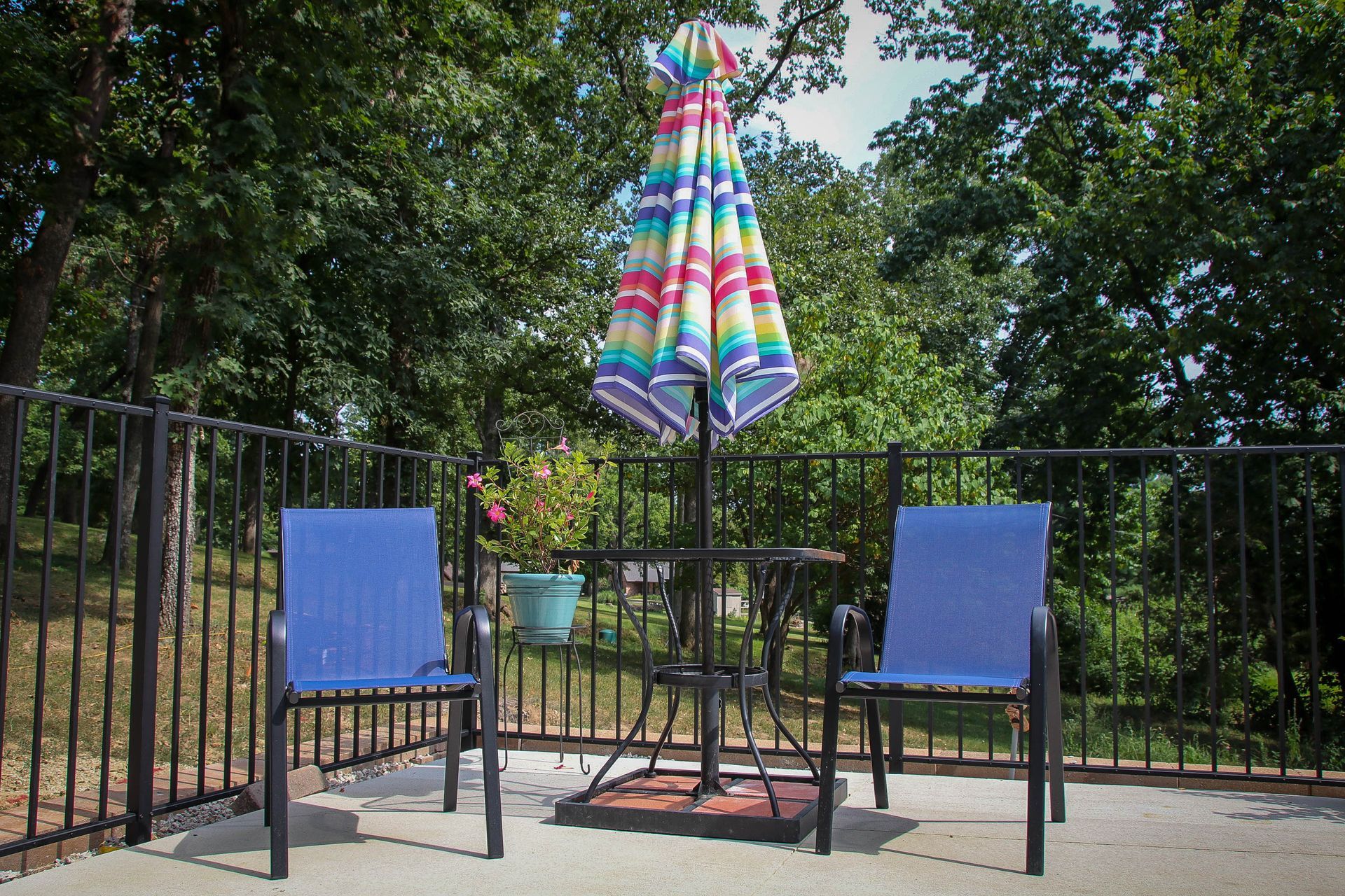 Two blue chairs and a table with umbrella on a patio overlooking trees.