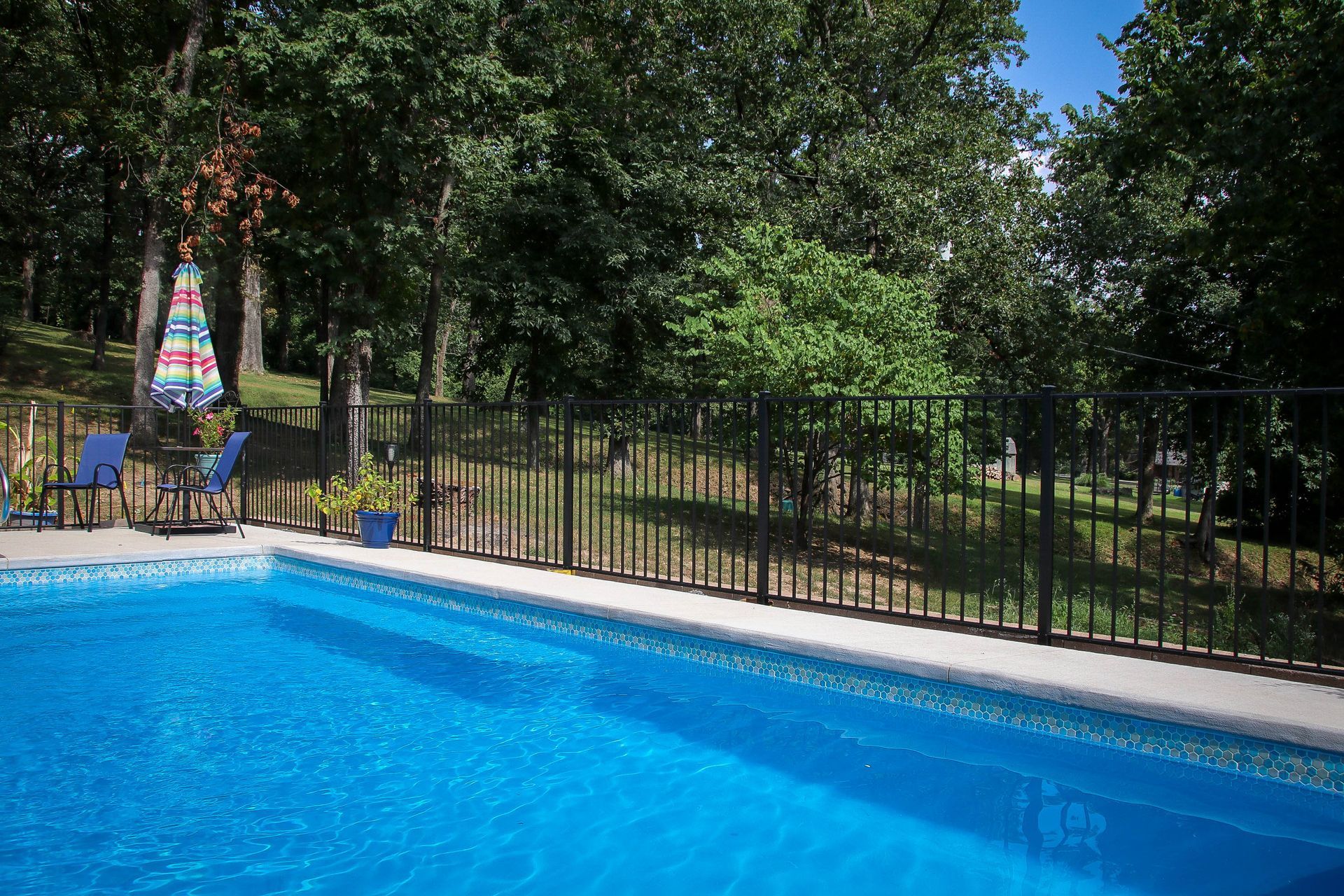 Poolside scene with blue water, black fence, chairs, umbrella, and trees.