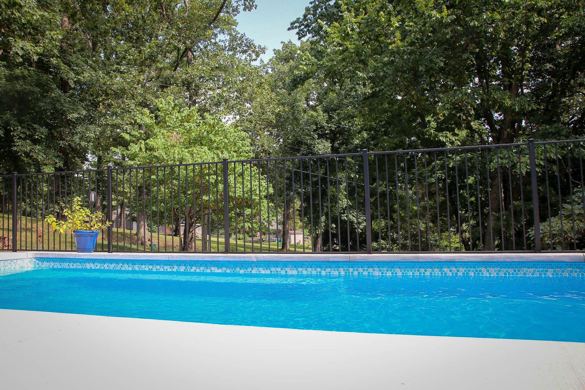 Pool with blue water and white coping, black fence, and trees in the background.