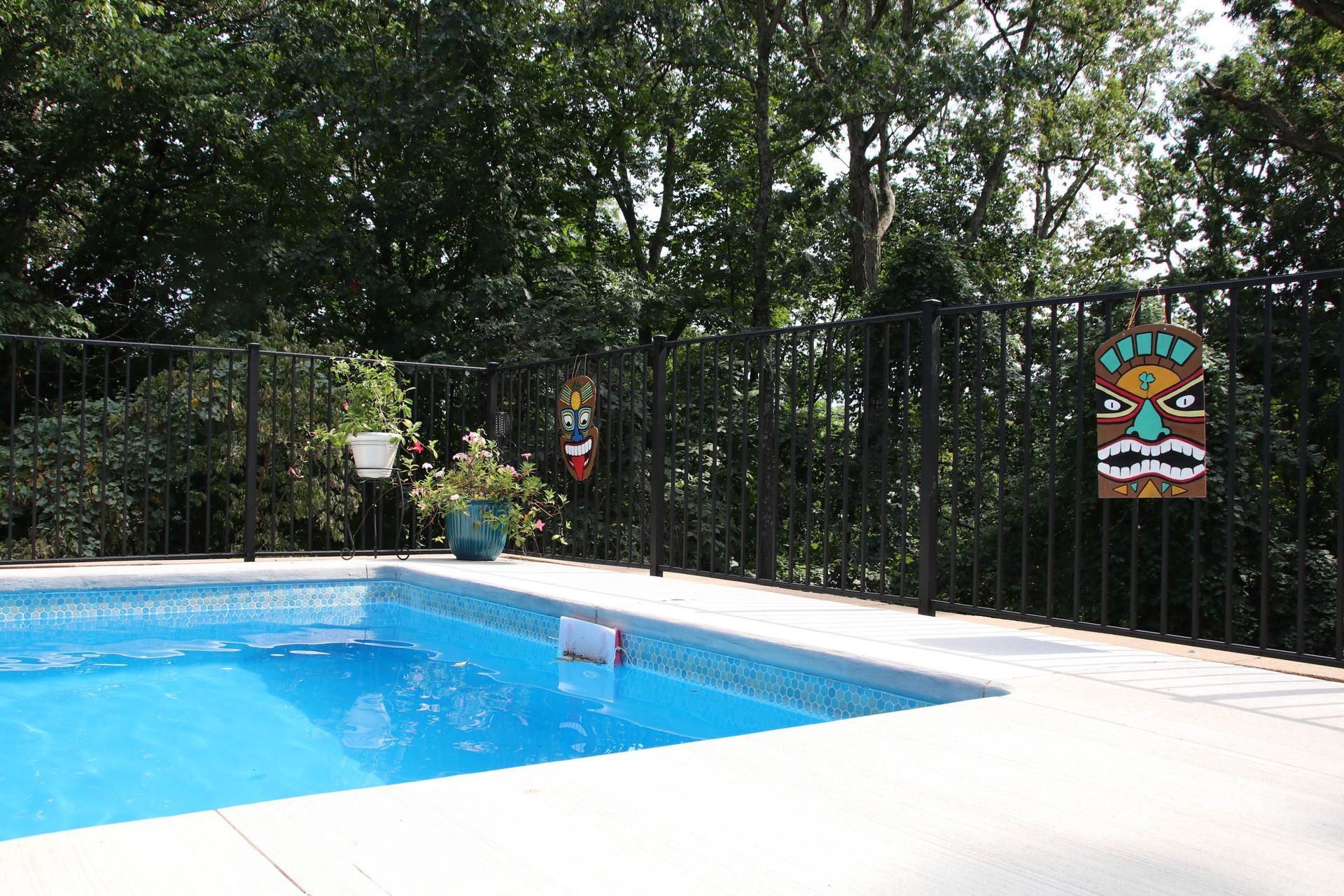 A blue swimming pool with a white concrete deck, black fence, and tiki decorations against a backdrop of trees.