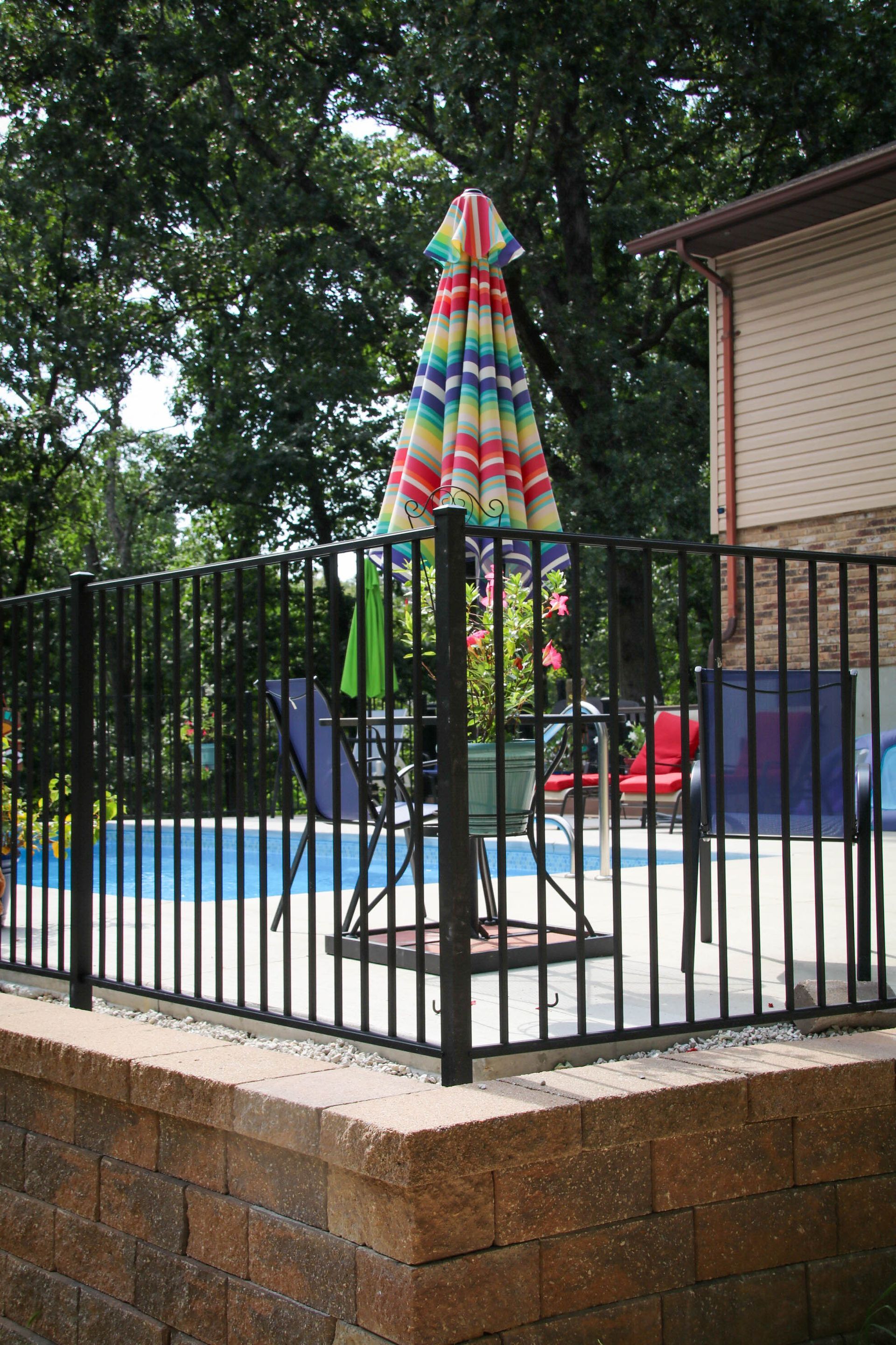 Black metal fence surrounds a pool with colorful umbrella and patio furniture.