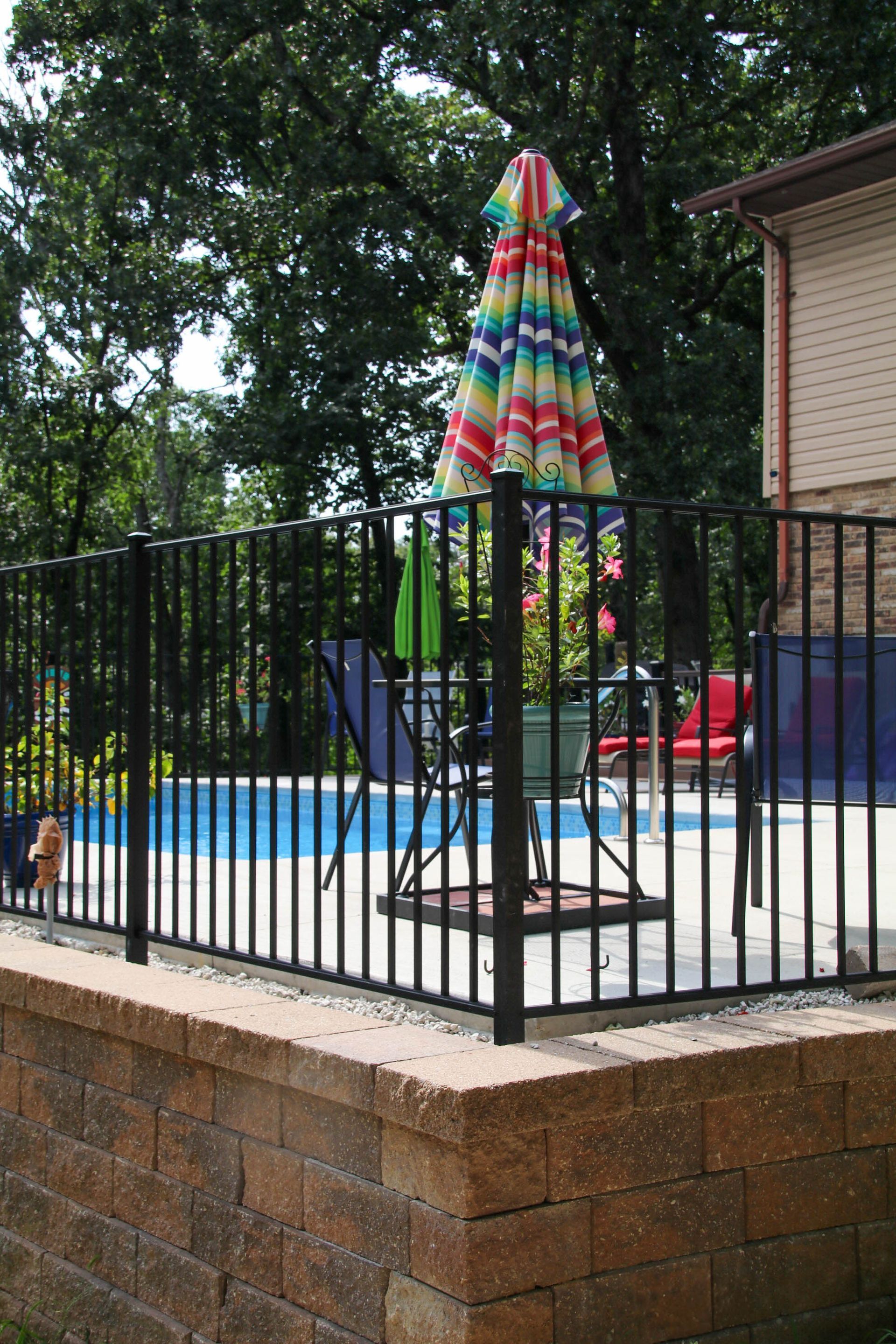 Black fenced pool area with colorful umbrella, red chair, and trees in background.