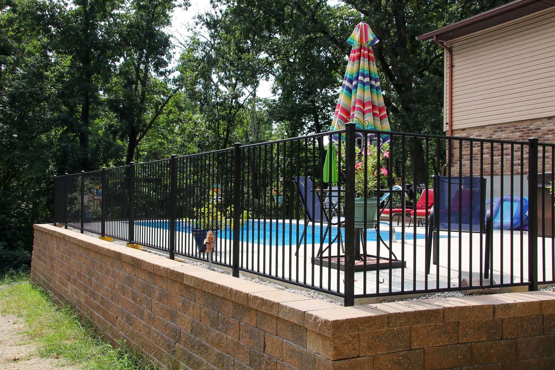 Black metal fence around a swimming pool built into a brick retaining wall; colorful umbrella in the background.