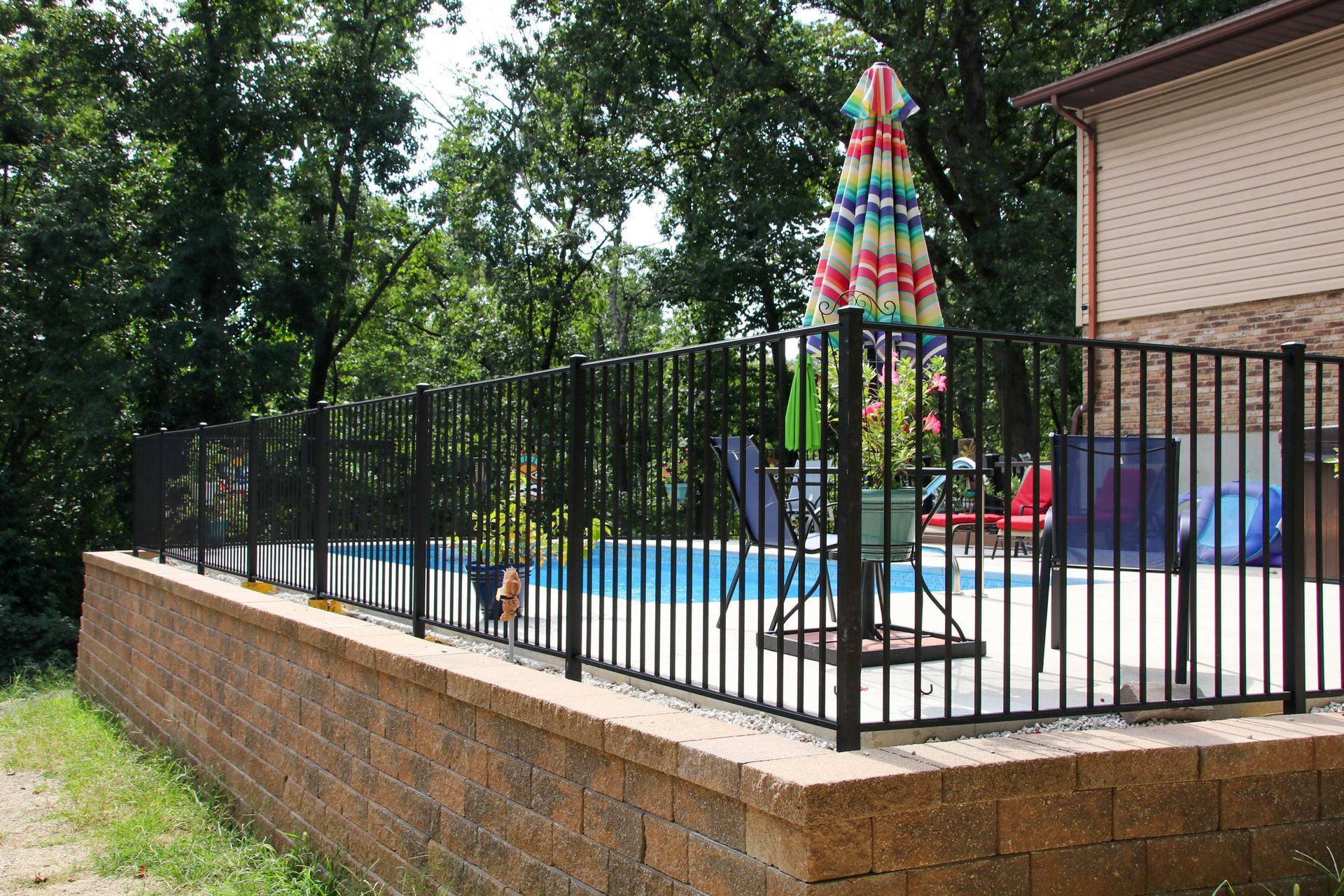 Black fence surrounds a swimming pool built into a brick retaining wall; umbrella and trees in background.